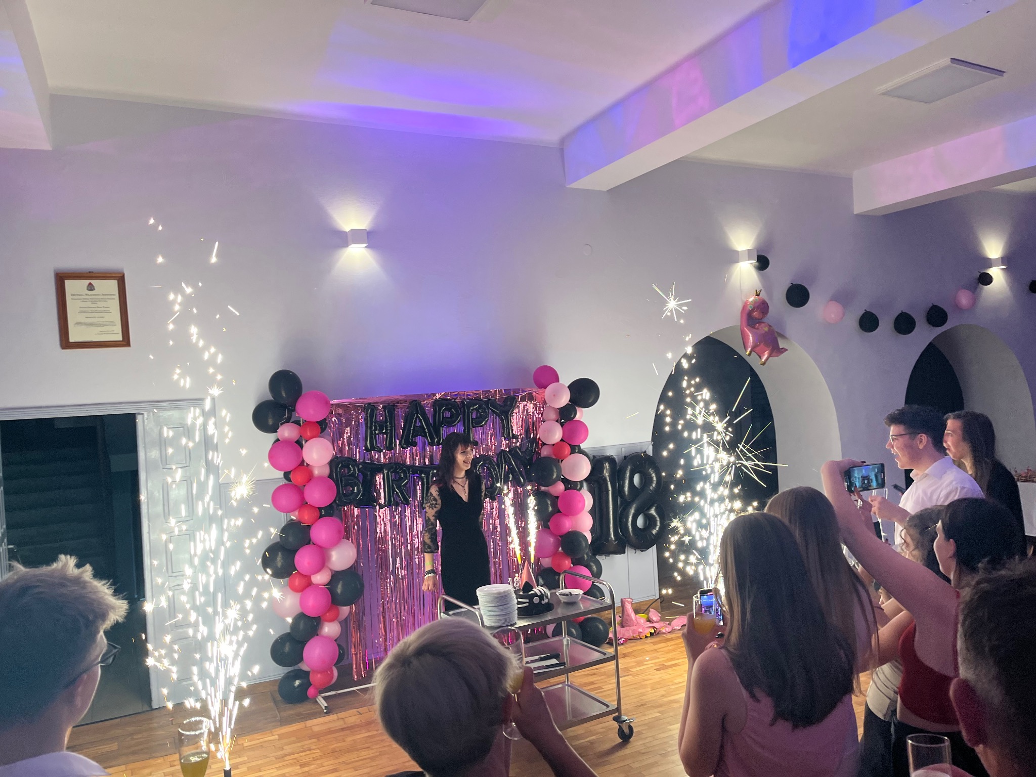 Uroczystość 18 urodzin z dekoracjami balonowymi, iskrzącymi fontannami i tortem na wózku. Goście robią zdjęcia telefonami, oświetlenie sali tworzy klimat.