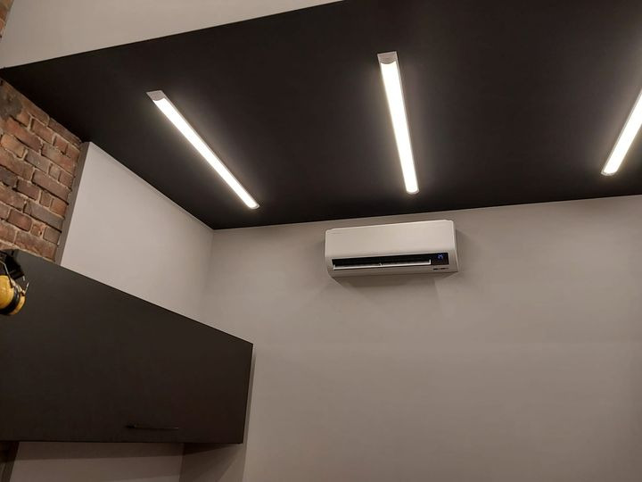 Biała jednostka klimatyzacyjna zamontowana na szarej ścianie pod trzema podłużnymi lampami LED na czarnym suficie, widoczny fragment ściany z cegły i szafka.