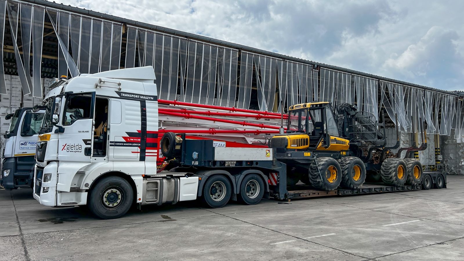 Ciężarówka z naczepą transportującą żółto-czarny kombajn leśny na tle magazynu z zasłonami paskowymi.