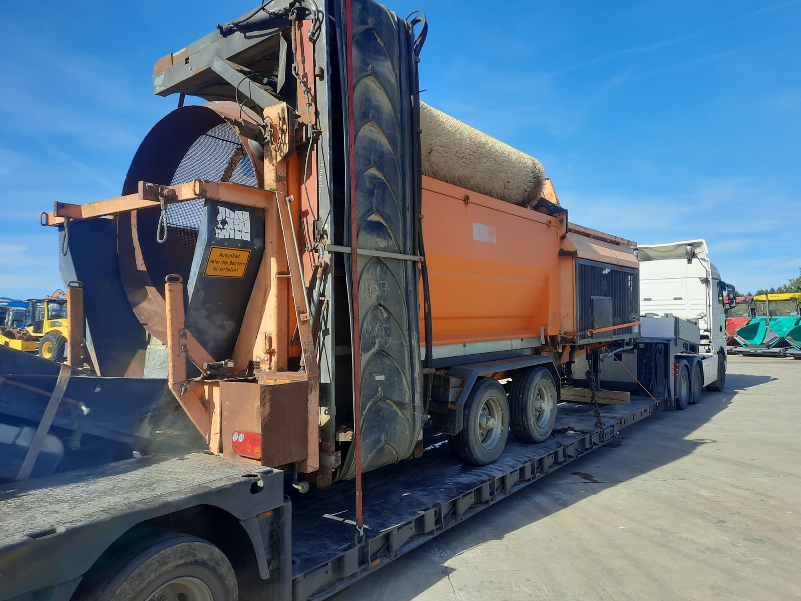 Pomarańczowy zestaw do przesiewania i transportu materiałów sypkich na niskopodwoziowej naczepie, widoczny znak ostrzegawczy w języku niemieckim.