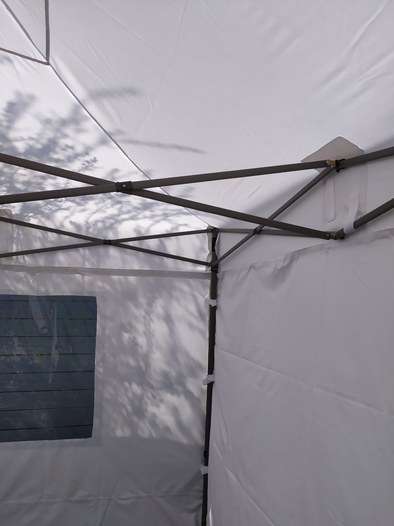 Wnętrze białego namiotu z widoczną metalową konstrukcją, fragmentem okna z żaluzjami i cieniem drzewa padającym na materiał ścianki.