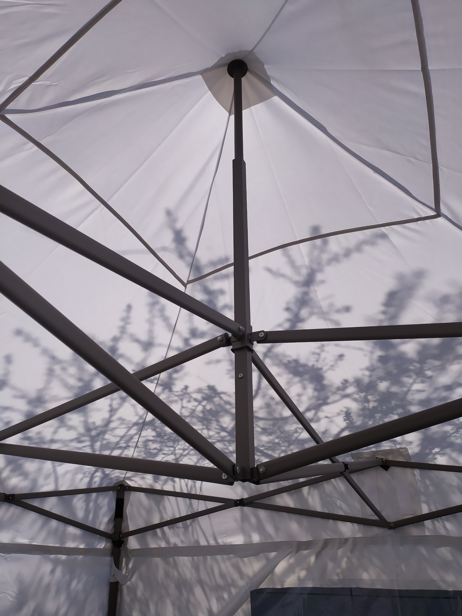 Wnętrze białego namiotu z metalową konstrukcją, widoczne cienie gałęzi drzew na materiale.