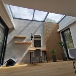 Nowoczesne wnętrze z oknami dachowymi z czarnymi ramami, betonową ścianą, drewnianą boazerią, biurkiem z lustrem, półkami i szarymi krzesłami.