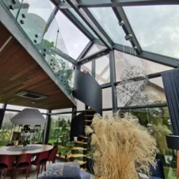 Nowoczesne wnętrze domu z przeszklonym dachem i schodami z balustradami ze szkła, widok na jadalnię i salon z elementami dekoracyjnymi, takimi jak lampy wiszące i trawa ozdobna.