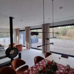 Nowoczesny salon z dużymi oknami, czarnym kominkiem wiszącym i minimalistycznym oświetleniem, widok na zewnątrz z drzewami i piaskiem, stół z dekoracją świąteczną.