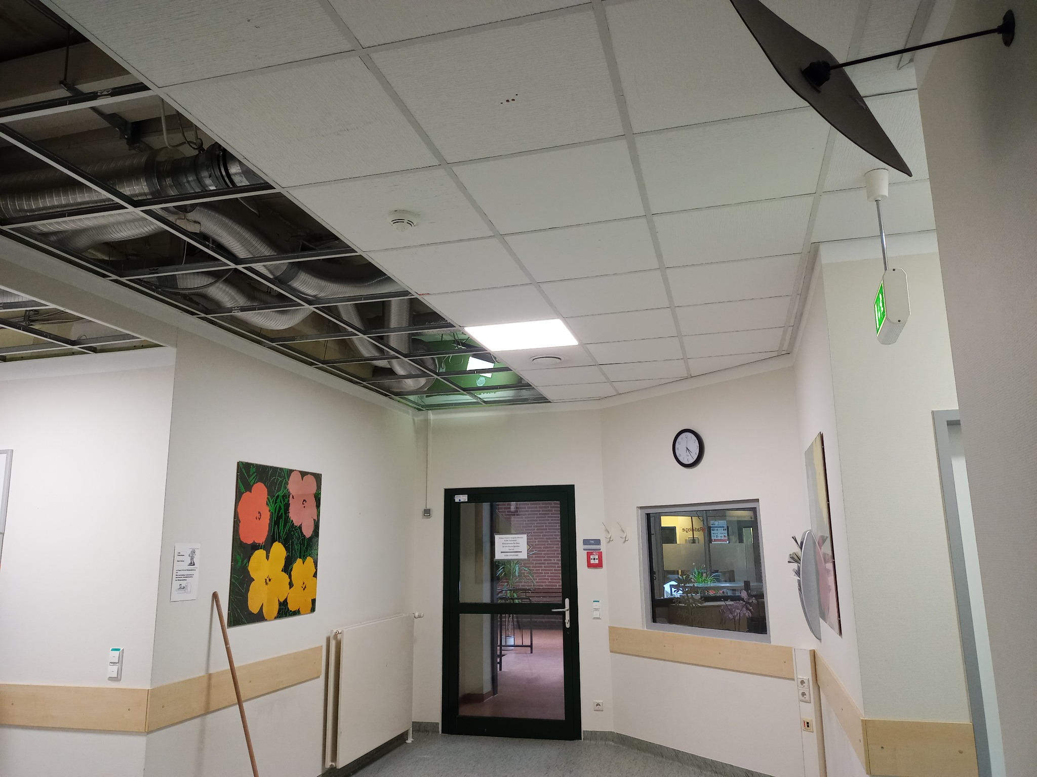 Widok na sufit podwieszany z odsłoniętą instalacją wentylacyjną, fragmentem oświetlenia i czujnikiem dymu w pomieszczeniu biurowym lub publicznym.