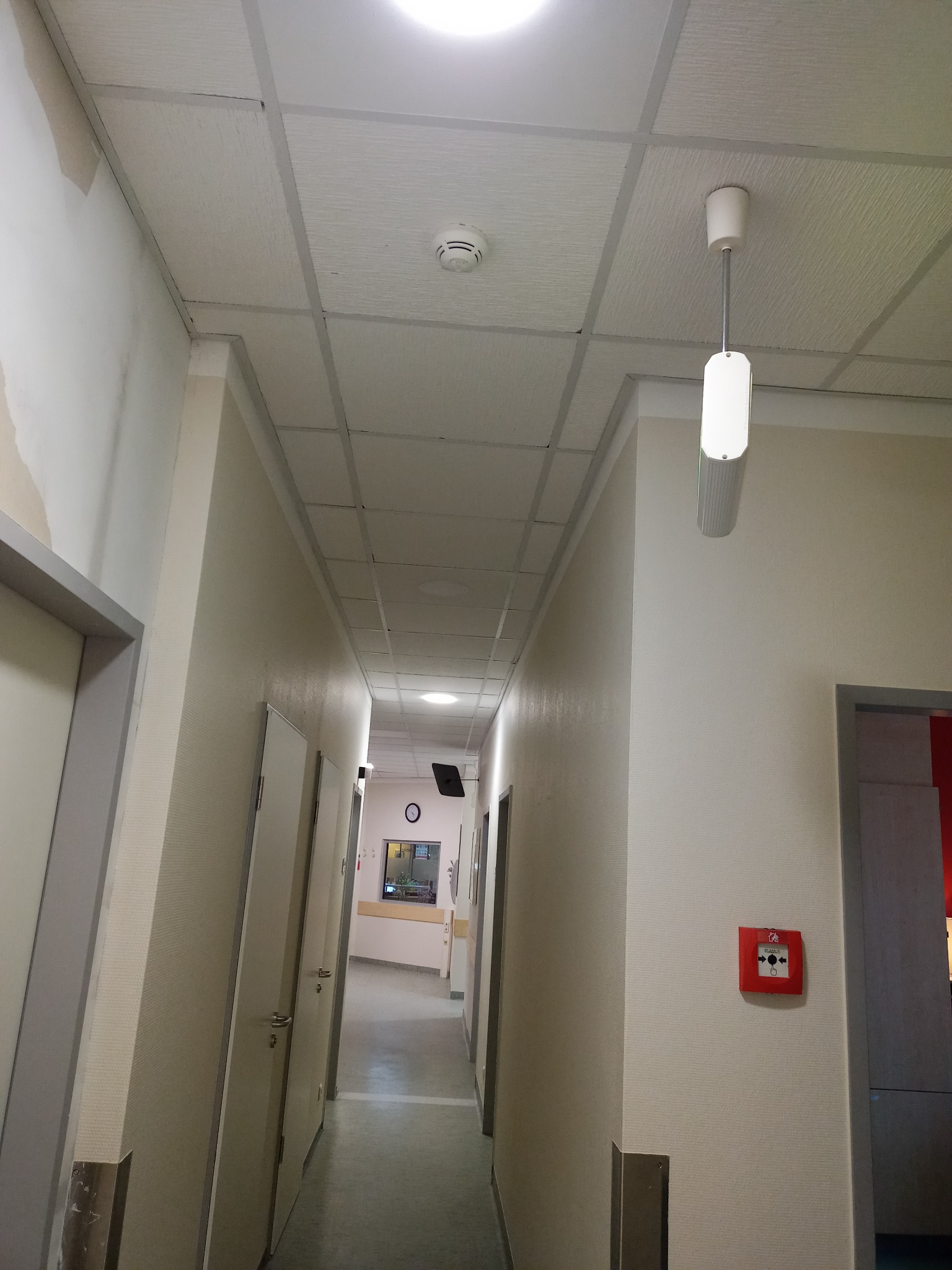 Korytarz biurowy z kasetonowym sufitem z widocznymi śladami po zalaniu na ścianie, oświetlony lampą wiszącą i okrągłym czujnikiem dymu.