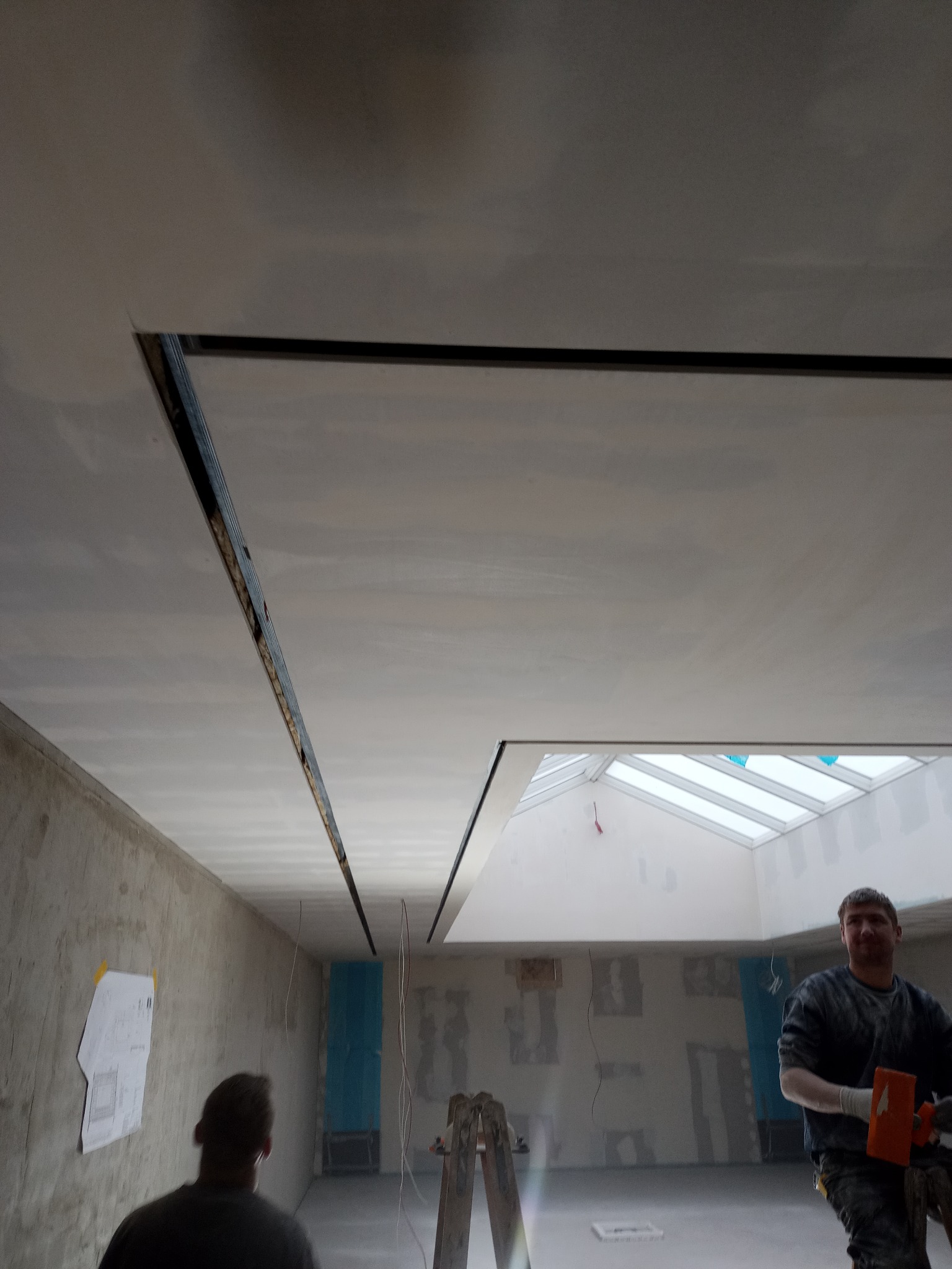 Wnętrze w trakcie remontu; widoczny sufit z zamontowanymi profilami, częściowo pokryty szpachlą, oraz okno dachowe; na ścianie wisi plan; pracownik zacierający szpachlę.