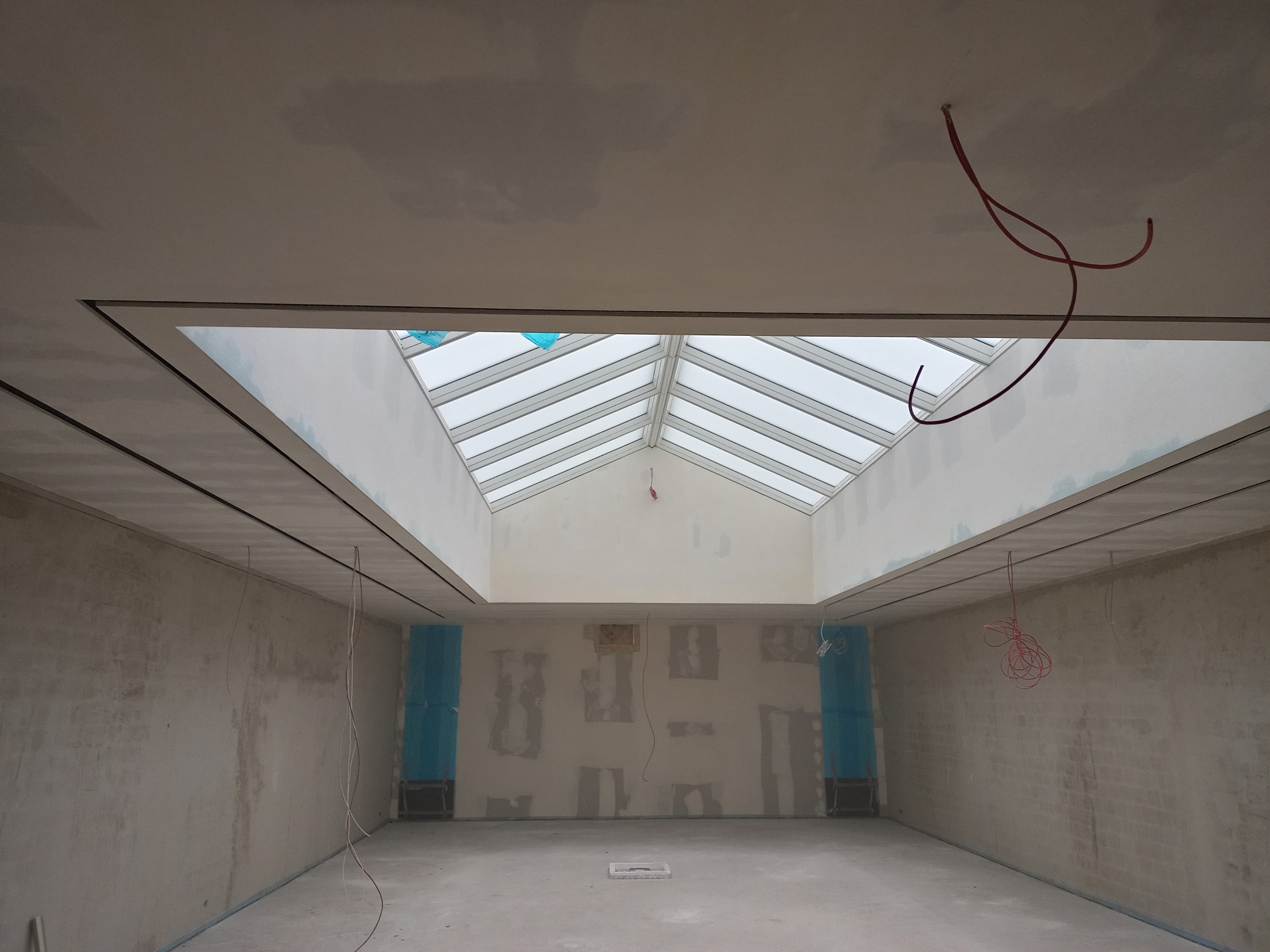 Wnętrze w trakcie remontu z widocznym świetlikiem dachowym, ściany przygotowane do wykończenia, zabezpieczone elementy instalacji, surowe wykończenie.