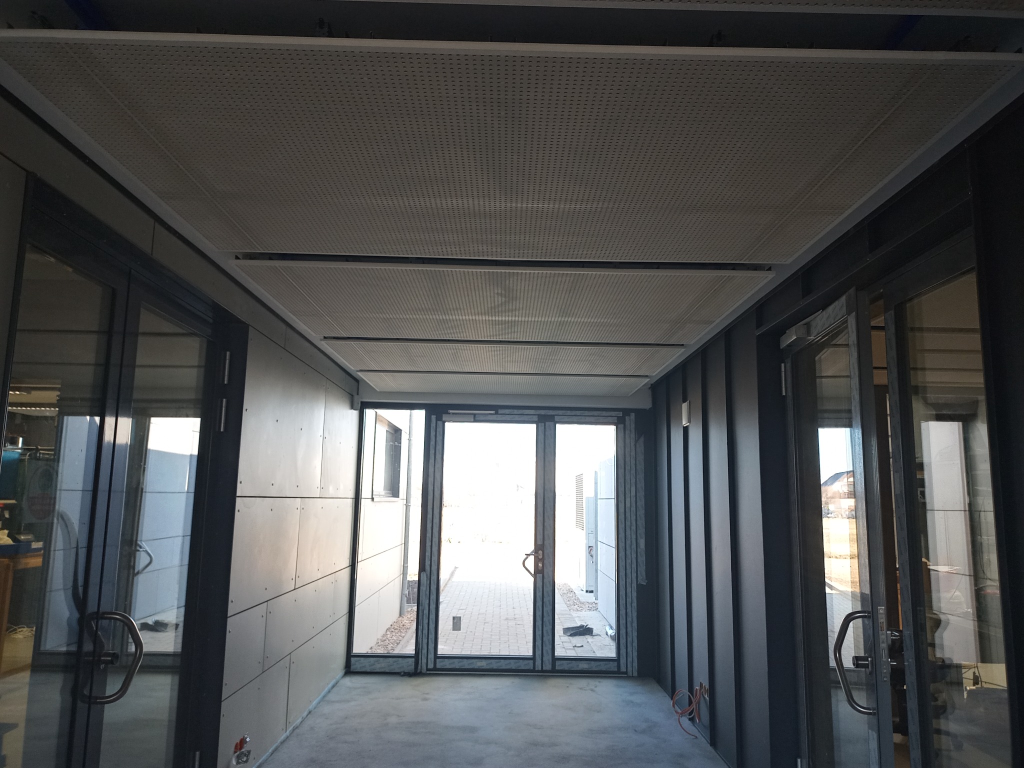 Korytarz z sufitami podwieszanymi z perforowanych paneli, ściany wykończone płytami, przeszklone drzwi wejściowe na końcu korytarza.