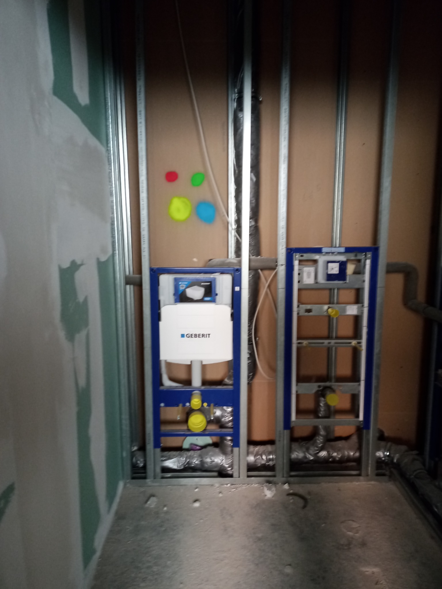 Stelaż podtynkowy Geberit zamontowany w konstrukcji z profili stalowych, częściowo zakryty płytą karton gips, widoczne kolorowe plamy na ścianie.