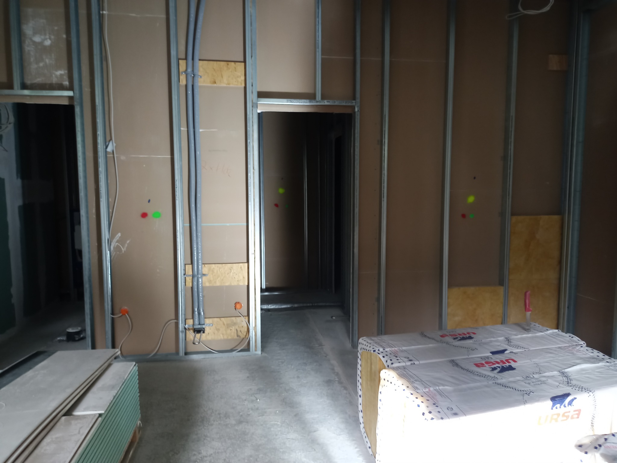 Wnętrze w trakcie prac wykończeniowych: metalowy stelaż, płyty, instalacje elektryczne, paczka wełny mineralnej URSA, widok na korytarz.