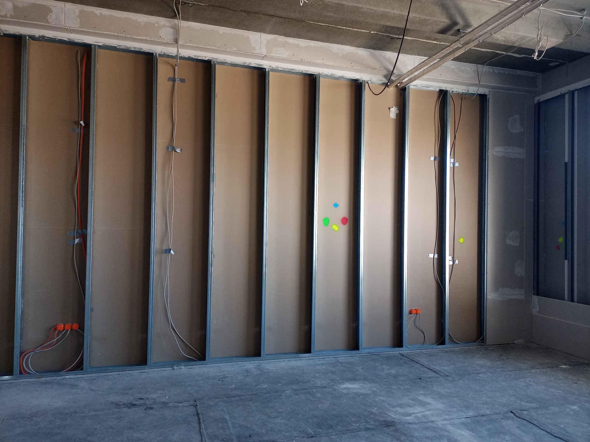 Montaż ścian działowych: metalowa konstrukcja z przytwierdzonymi płytami, widoczne przewody elektryczne w peszlach i puszki instalacyjne, surowy betonowy sufit, szare podłoże.