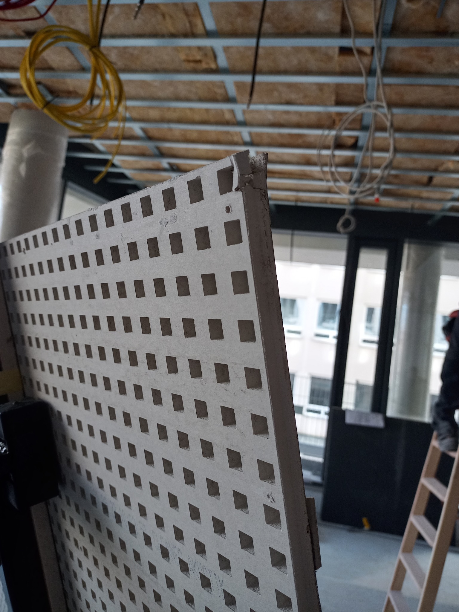 Perforowana płyta gipsowo-kartonowa z widoczną konstrukcją sufitu podwieszanego i instalacją elektryczną w tle, drabina w pomieszczeniu w trakcie remontu.