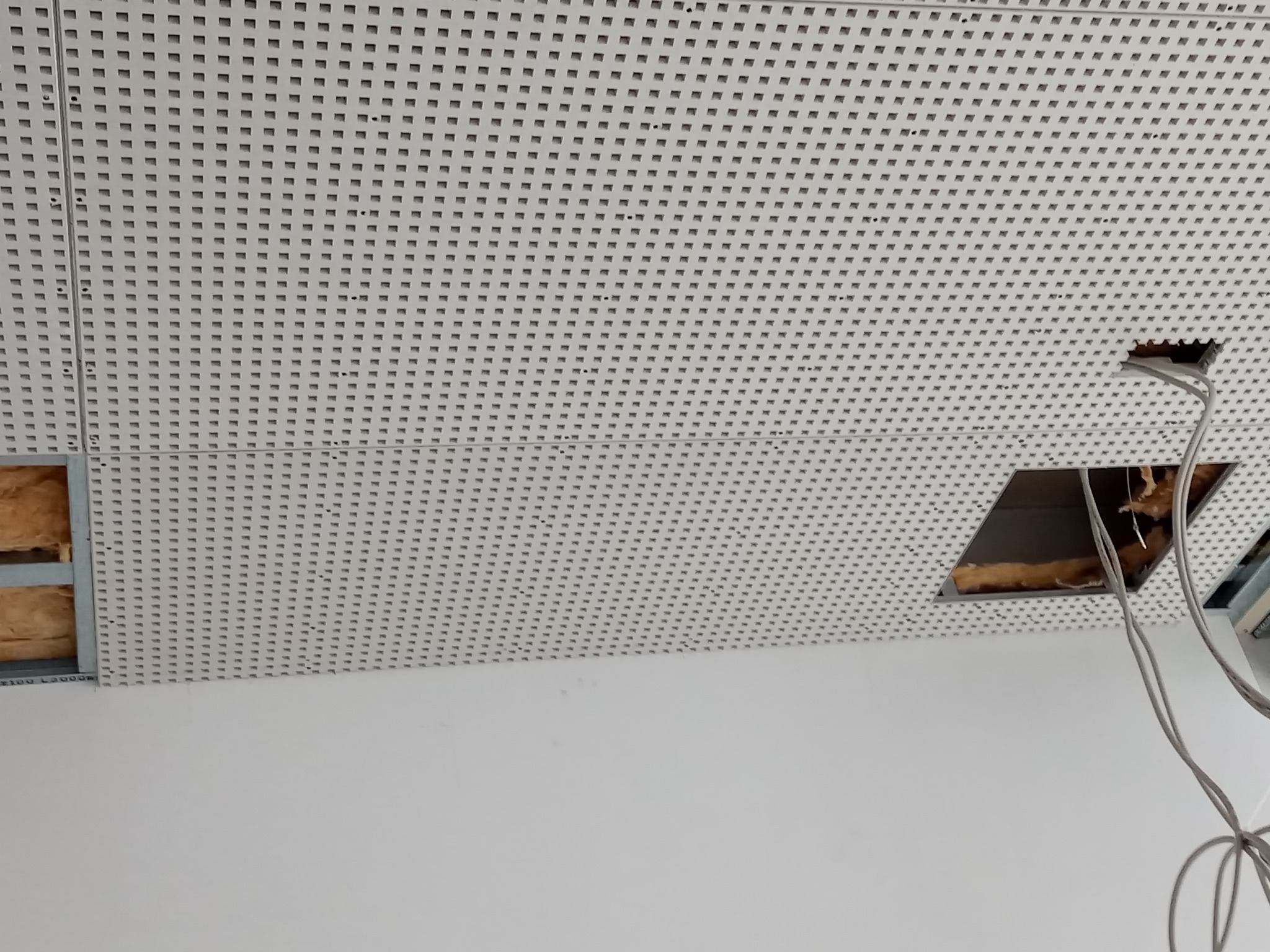 Perforowany sufit podwieszany z widoczną izolacją i przewodami w otworach montażowych.