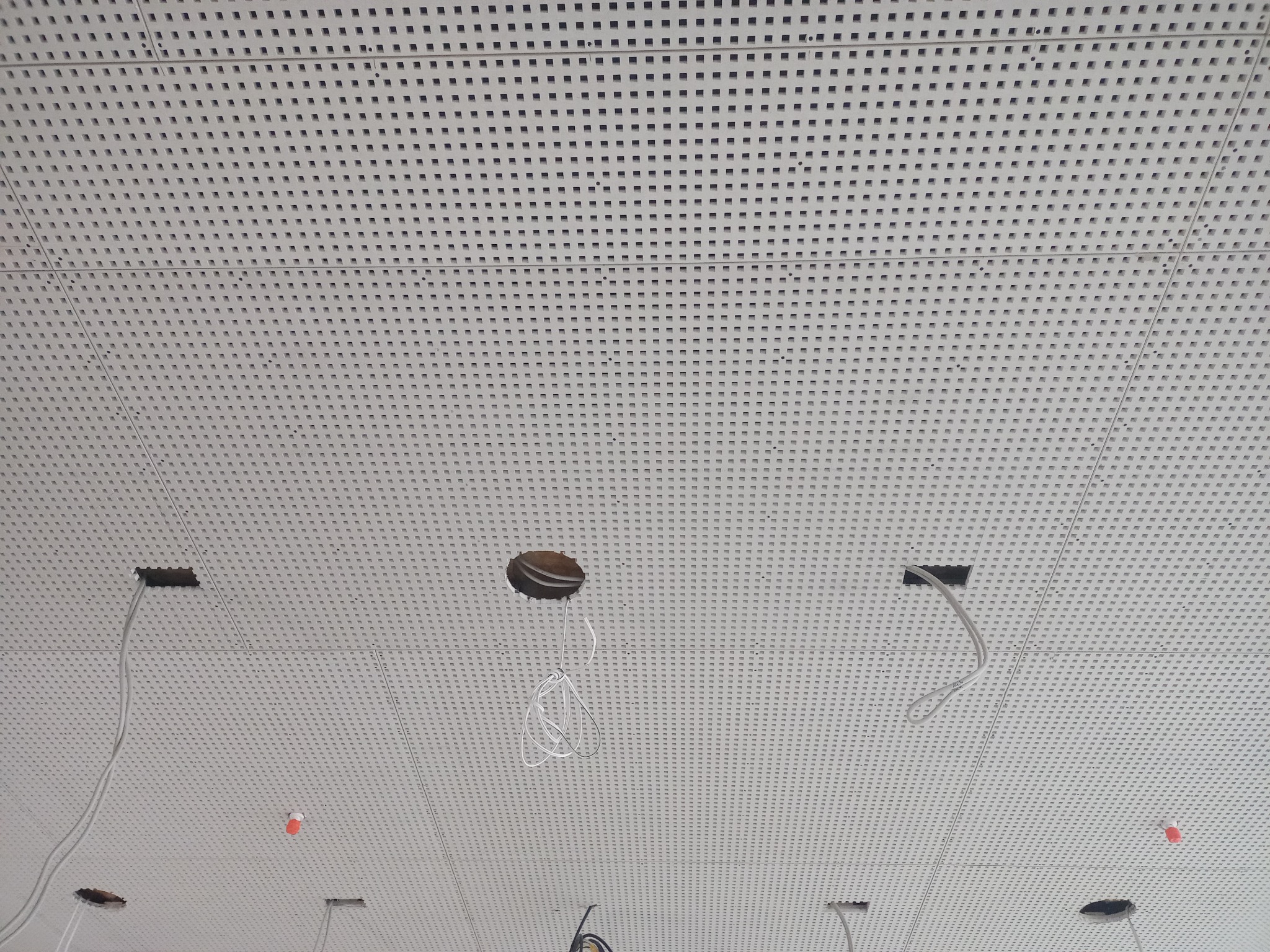 Perforowany sufit podwieszany z otworami na instalacje elektryczne i wystającymi przewodami, widoczne pomarańczowe znaczniki.