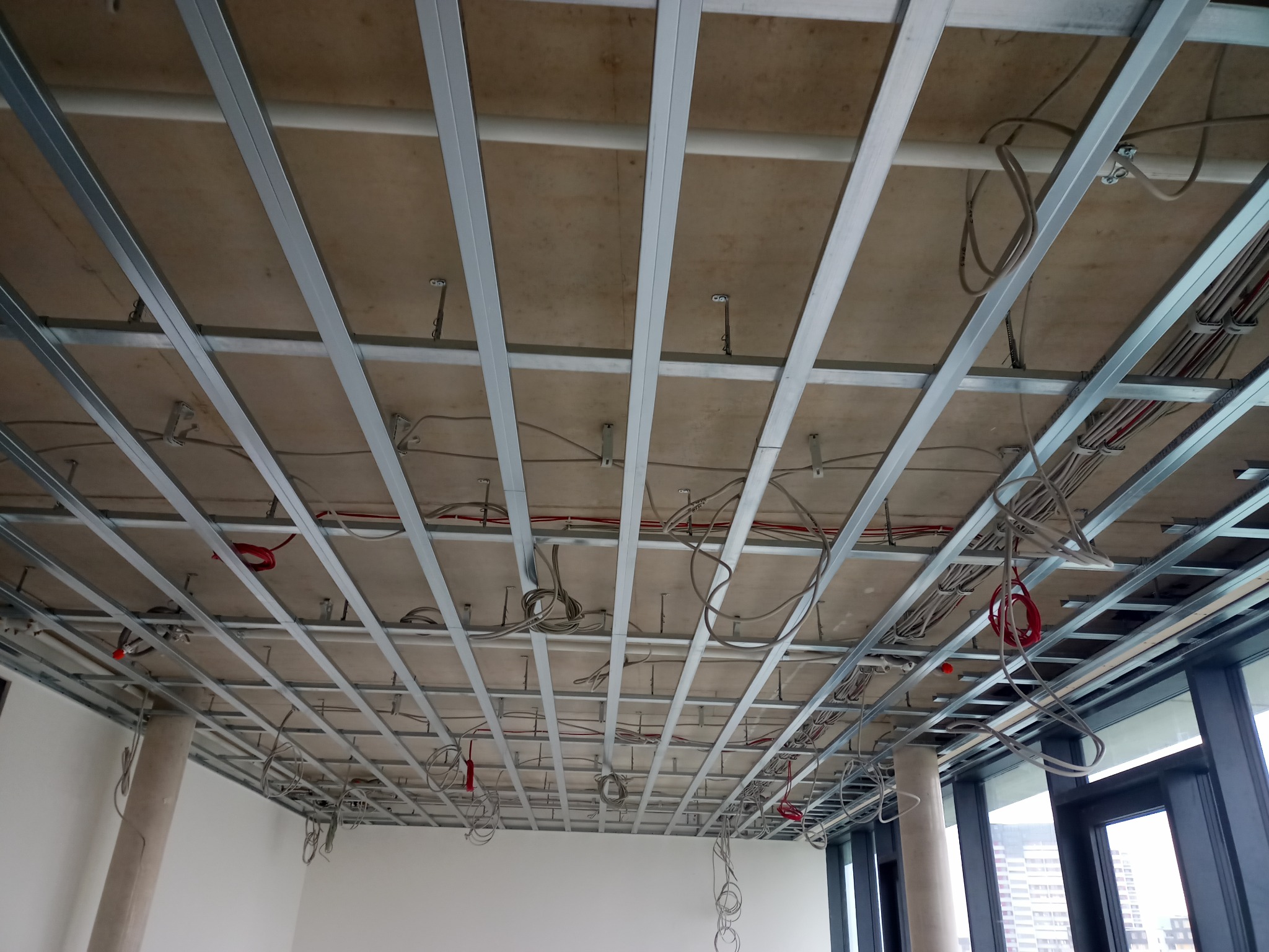 Metalowa konstrukcja podwieszanego sufitu z widocznymi przewodami elektrycznymi, rurami i elementami mocującymi na tle surowego stropu w nowym budynku z dużymi oknami.