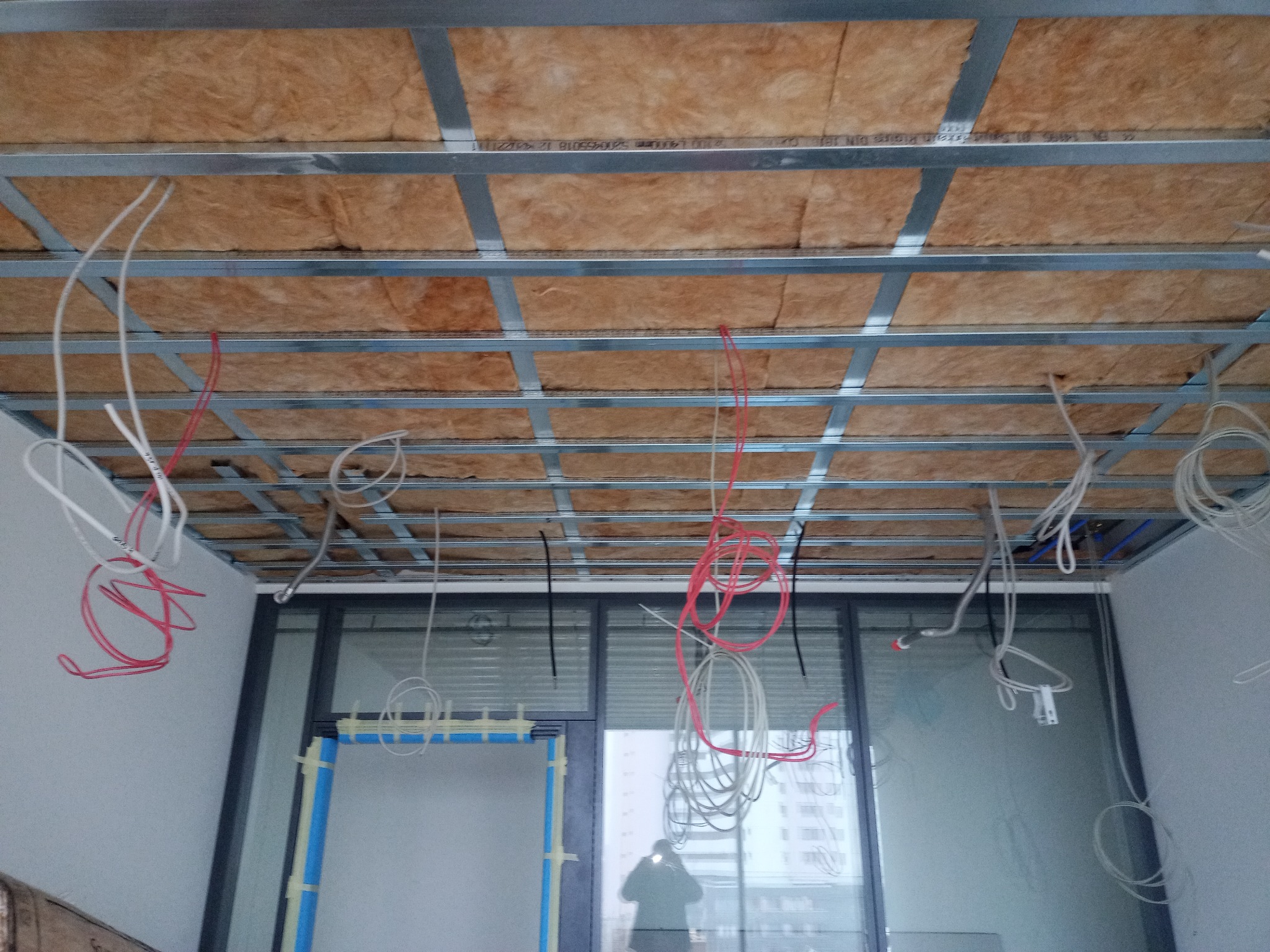 Montaż sufitu podwieszanego: widoczne profile metalowe, izolacja z wełny mineralnej i zwisające kable elektryczne w nowym budynku.