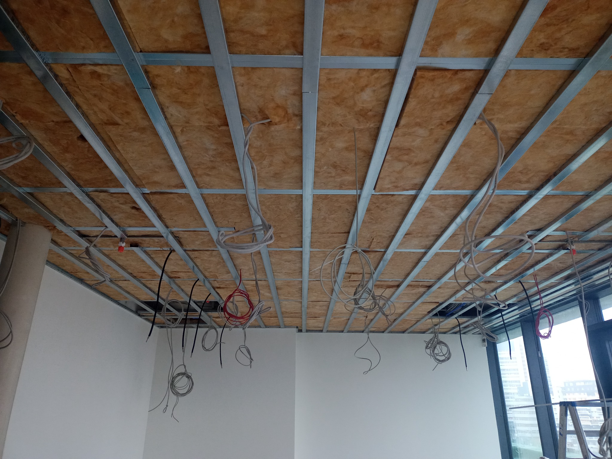 Metalowa konstrukcja podwieszanego sufitu z widoczną izolacją z wełny mineralnej i zwisającymi przewodami elektrycznymi w nowym pomieszczeniu z oknem.