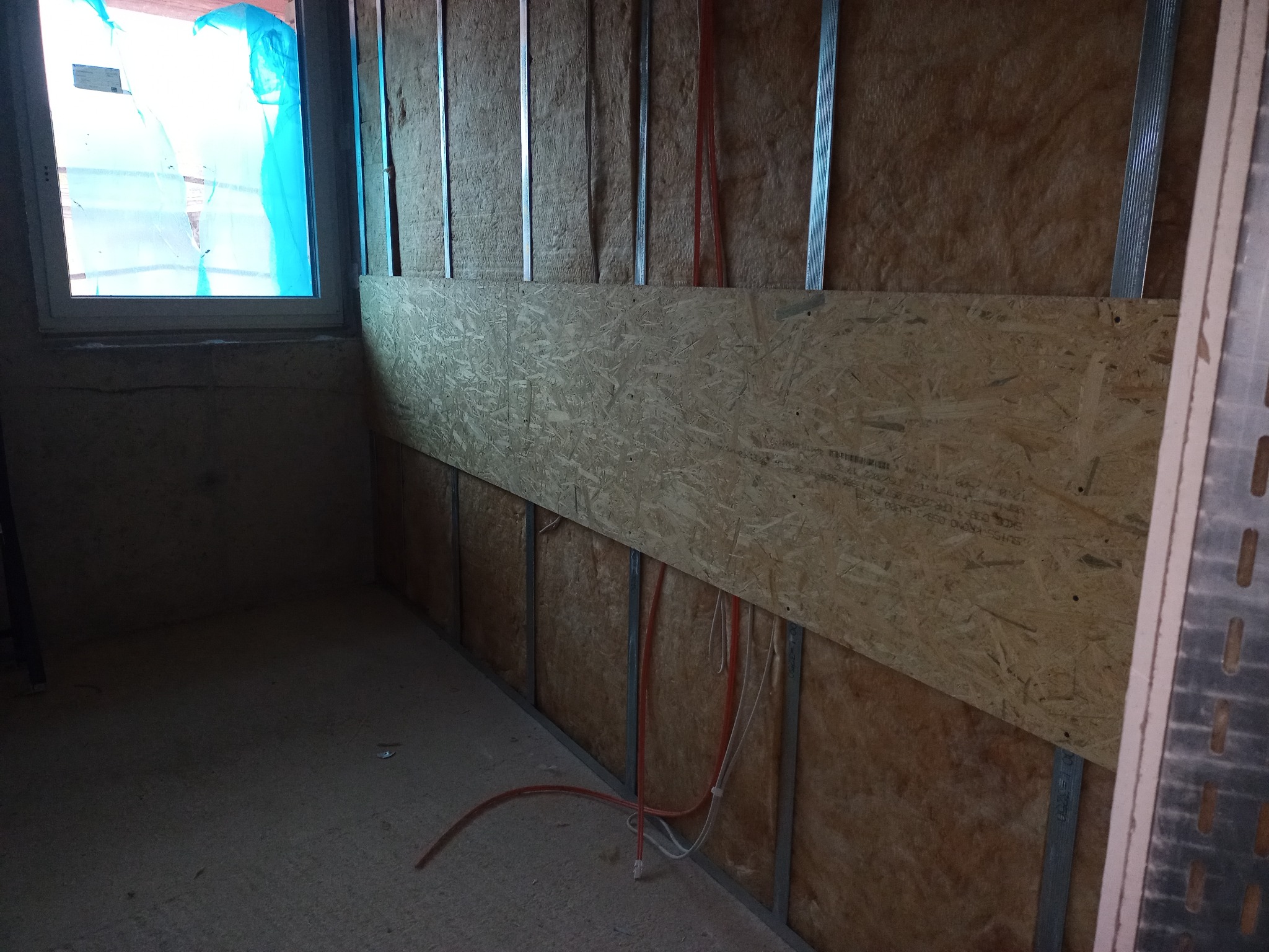 Wnętrze w trakcie remontu: ściana z izolacją z wełny mineralnej, metalowe profile, płyta OSB, instalacja elektryczna i okno z niebieską folią ochronną.