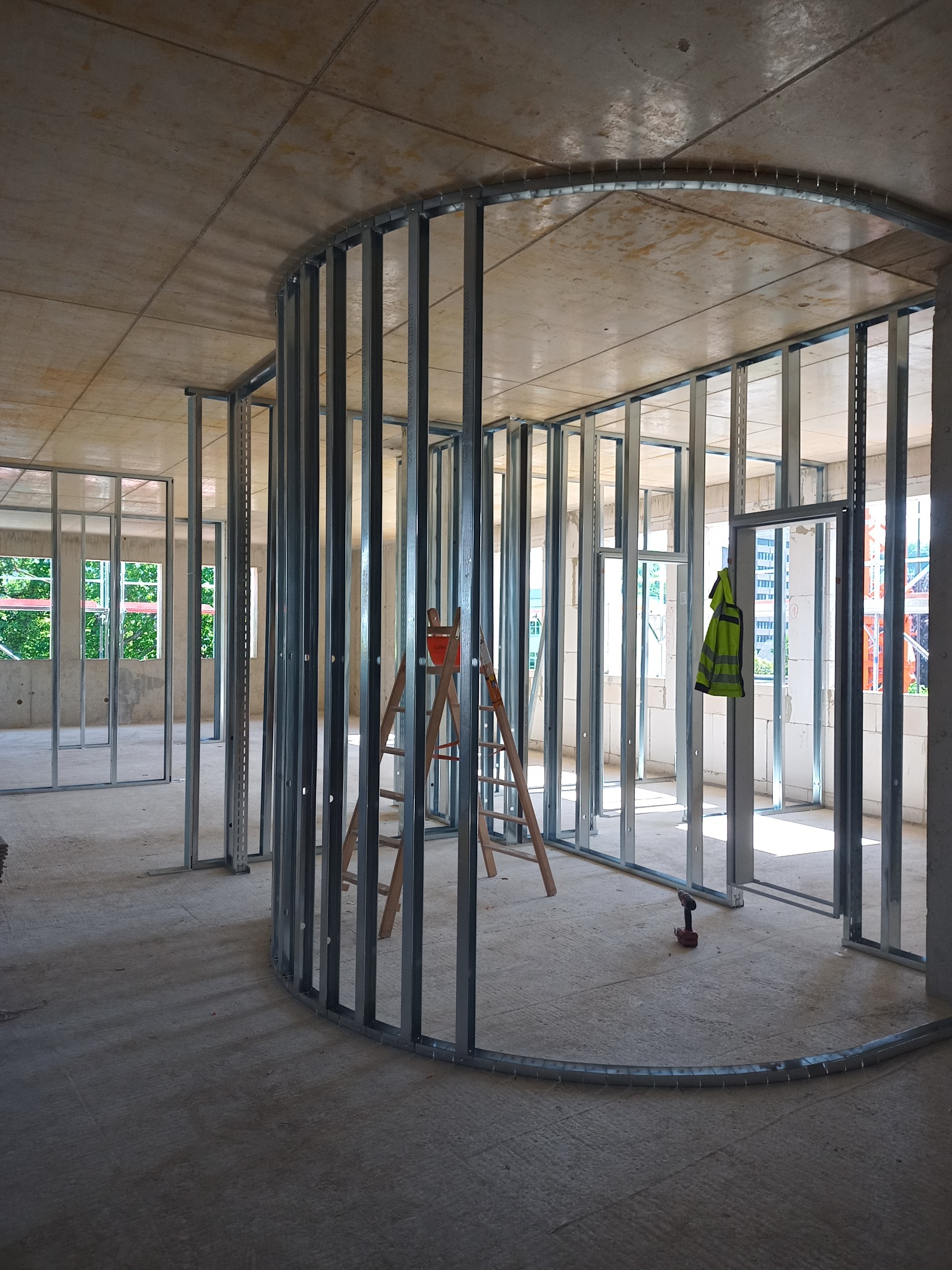 Stelaż z profili metalowych tworzący zaokrągloną ścianę działową w surowym stanie, widoczne narzędzia i drabina, żółta kamizelka odblaskowa w tle.