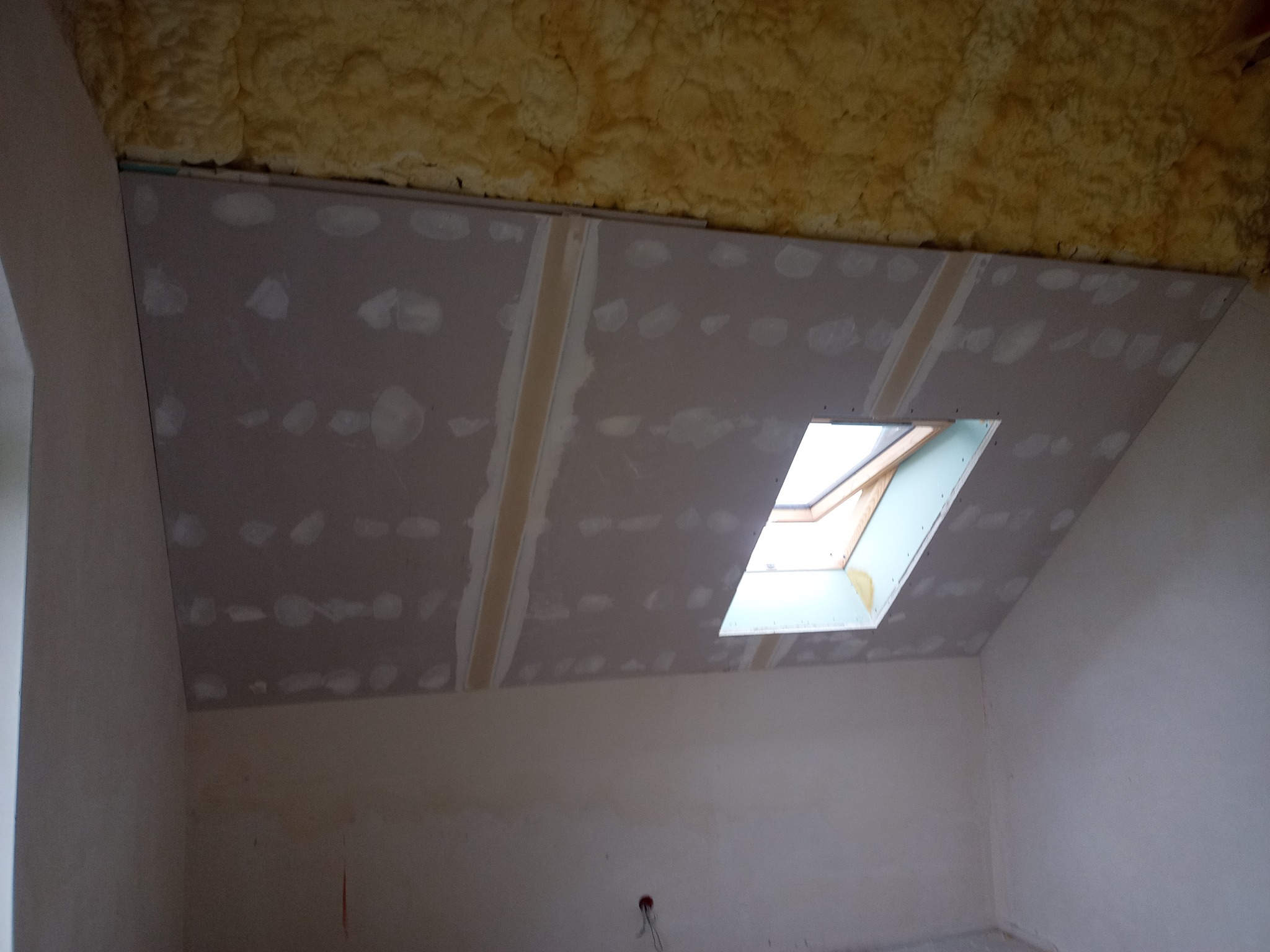 Poddasze w trakcie wykańczania: zamontowana płyta kartonowo-gipsowa na skośnym suficie z oknem dachowym, widoczne łączenia płyt przed szpachlowaniem, izolacja z pianki na poddaszu.