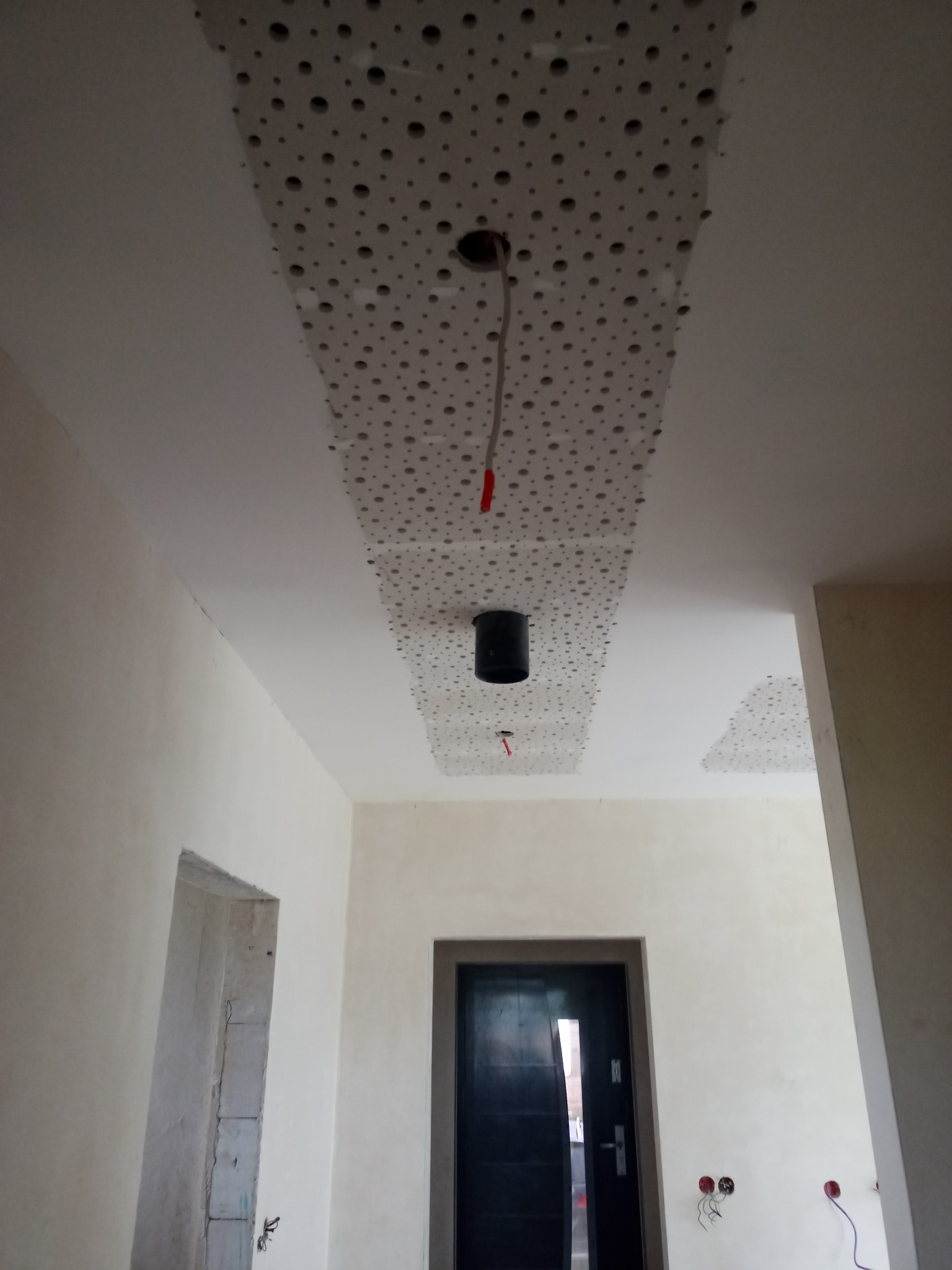 Fragment sufitu z widocznymi panelami akustycznymi z otworami, częściowo zamontowanymi, z wystającymi kablami do oświetlenia, w trakcie prac wykończeniowych wnętrza.