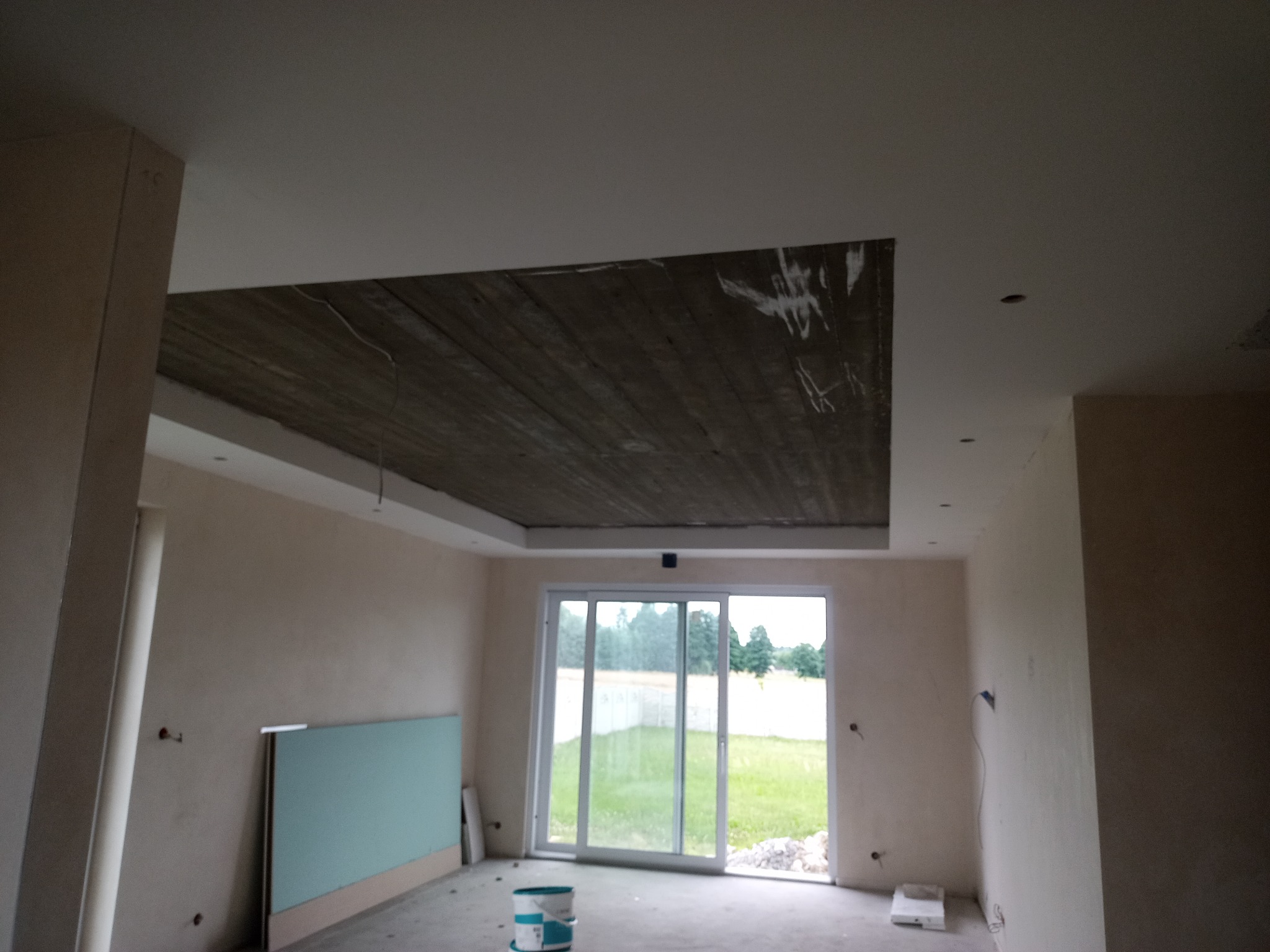Wnętrze w trakcie remontu: sufit z widoczną konstrukcją podwieszaną, ściany przygotowane do malowania, okno z widokiem na zieleń, oraz wiadro z farbą na podłodze.