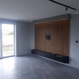 Nowoczesny salon z szarą podłogą imitującą marmur, ścianami w odcieniach szarości i drewnianą ścianką dekoracyjną z podświetleniem LED, pod którą znajduje się czarna szafka RTV.