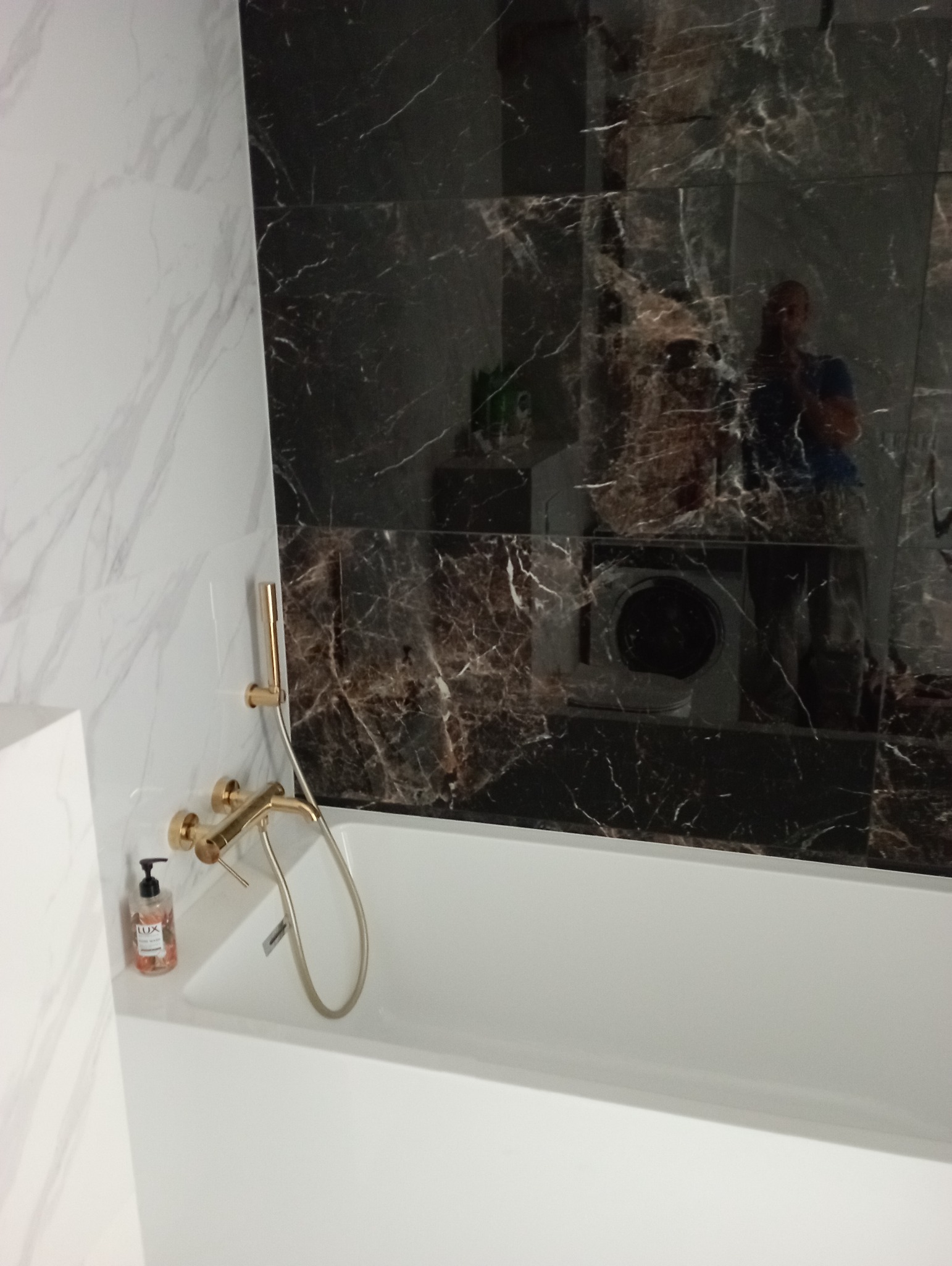 Biała wanna z złotą armaturą i dozownikiem mydła, obok ściana wyłożona płytkami imitującymi czarny marmur, w których odbija się pralka i osoba robiąca zdjęcie.