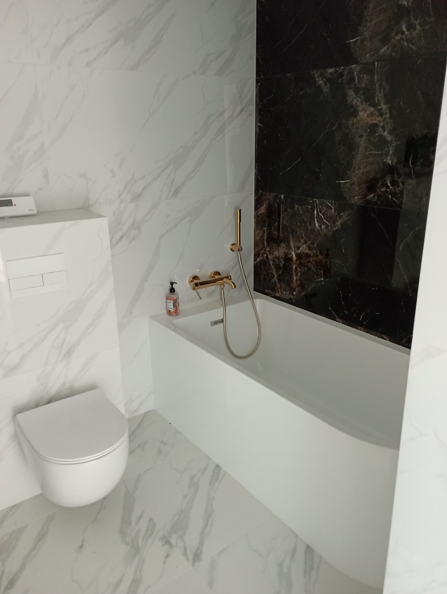 Nowoczesna łazienka z białą armaturą, złotą baterią wannową i okładziną z białego marmuru oraz czarną ścianą z motywem marmuru.