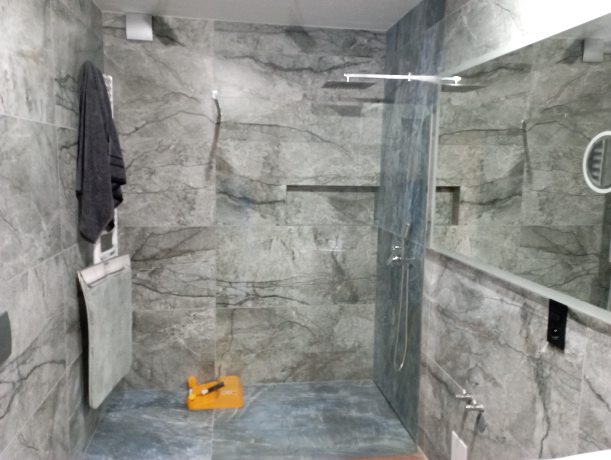 Wykończona łazienka z szarymi płytkami imitującymi marmur, deszczownicą i lustrem z podświetleniem. Na ścianie wisi szary ręcznik, a na podłodze leży żółta poziomica.