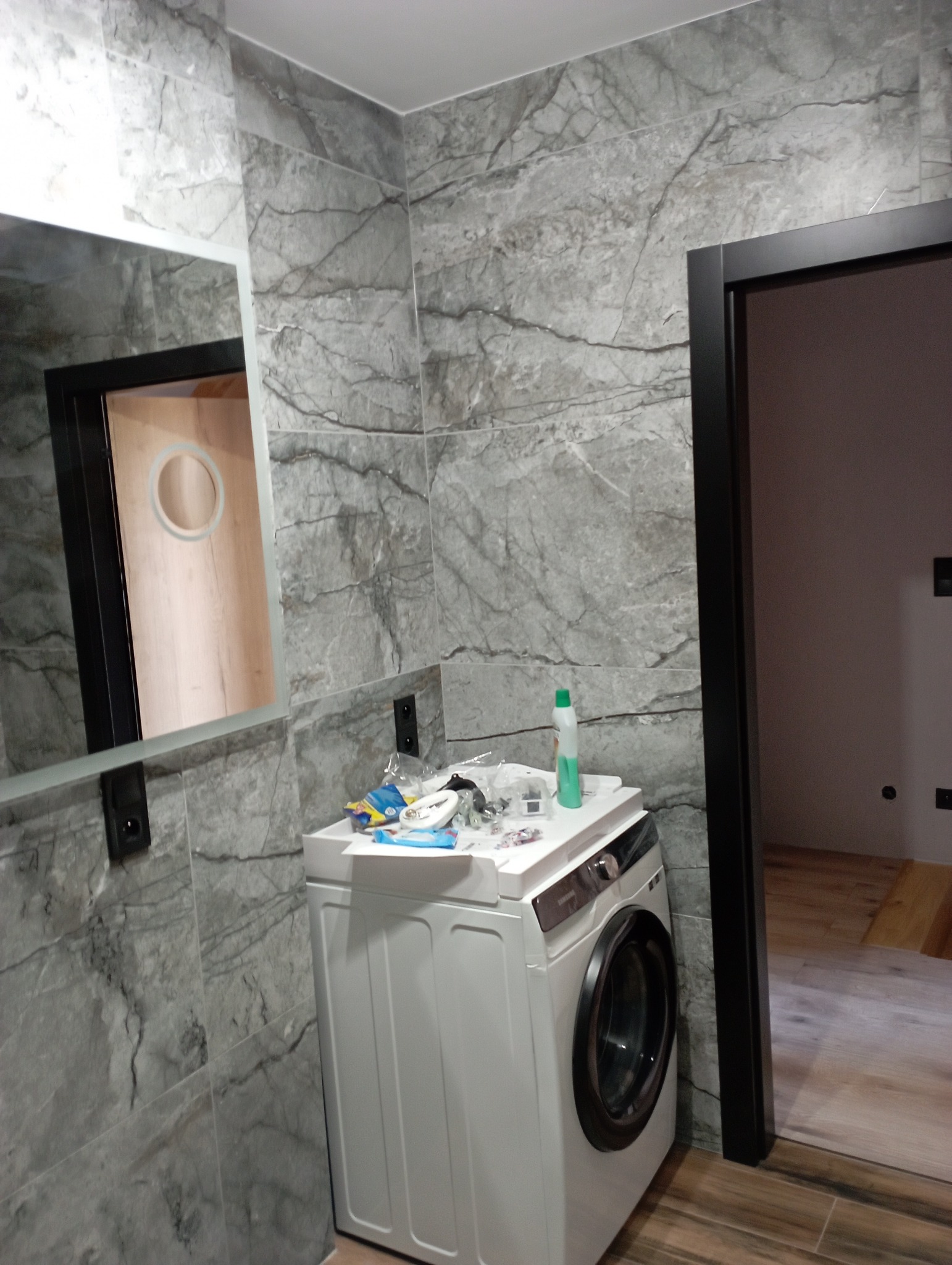 Łazienka w trakcie remontu: szare płytki imitujące marmur na ścianach, pralka z akcesoriami na górze, czarna rama lustra i drzwi, widoczna podłoga imitująca drewno.
