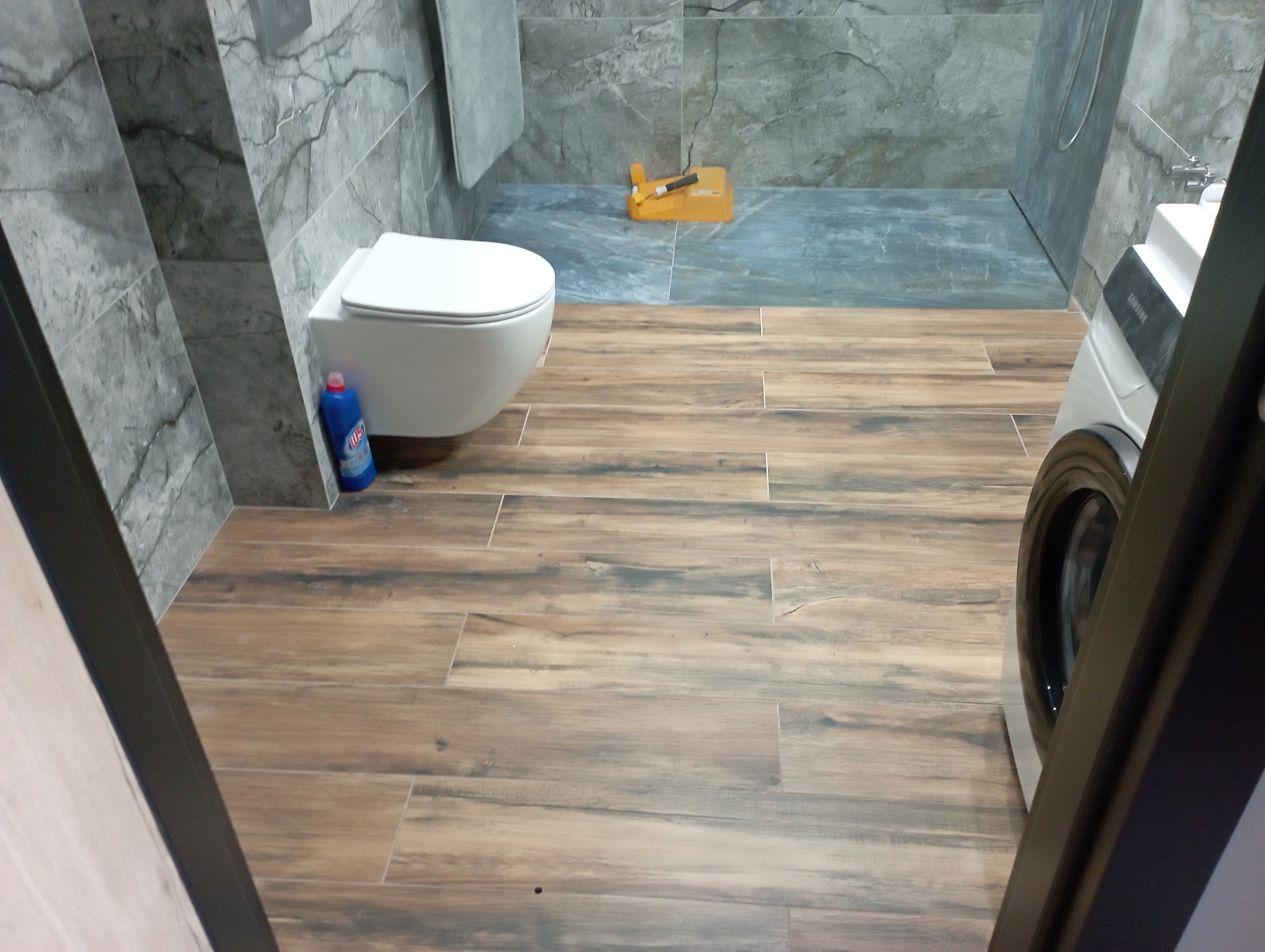 Ukończona łazienka z szarymi płytkami imitującymi marmur na ścianach, drewnopodobnymi płytkami na podłodze, wiszącą toaletą i pralką. Na podłodze leży żółta poziomica. Widoczna butelka płynu...