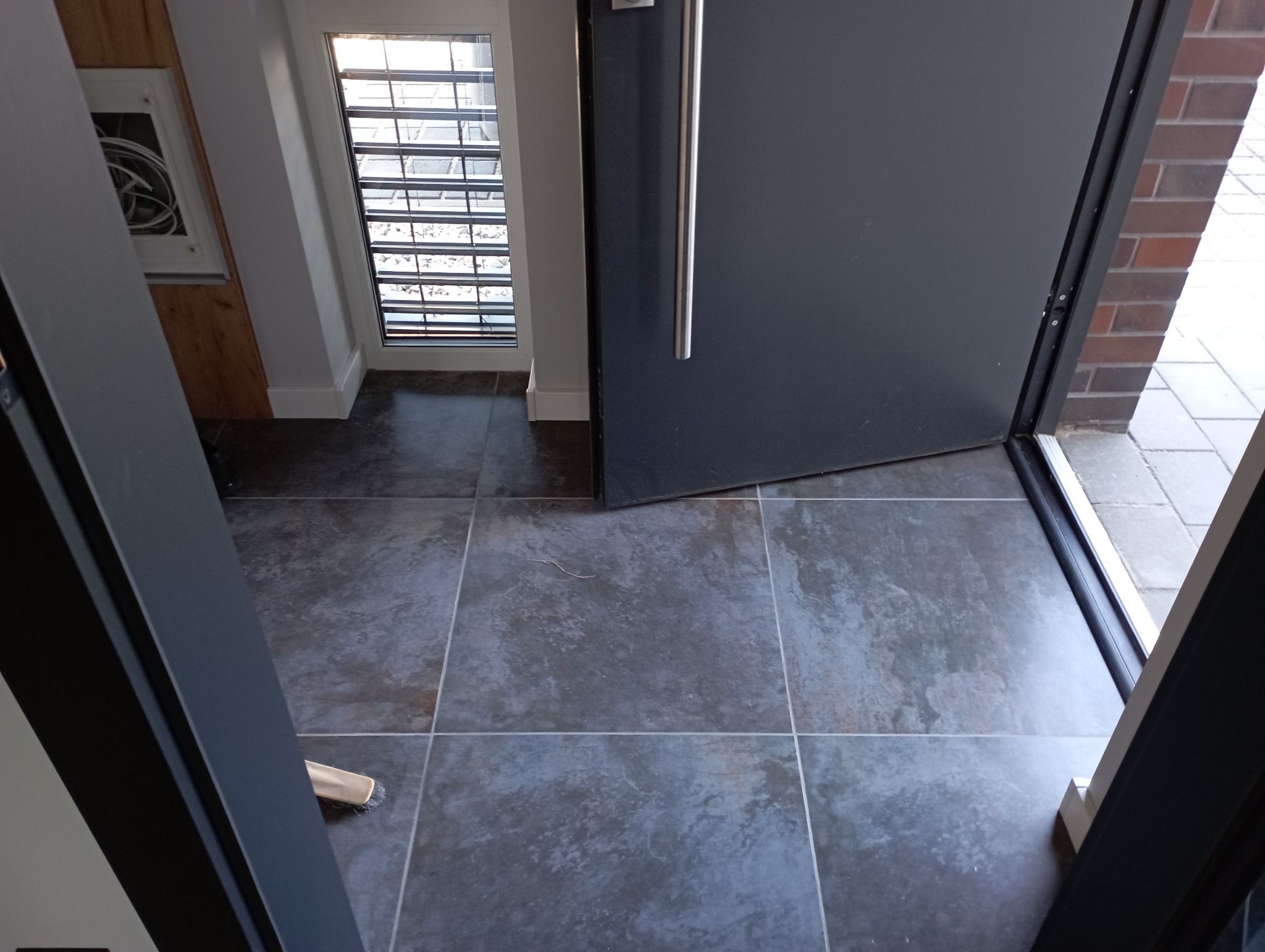 Ułożona szara płytka podłogowa w przedpokoju, widoczny fragment drzwi wejściowych z długim uchwytem oraz okno z żaluzjami w tle.