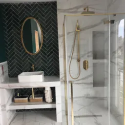 Nowoczesna łazienka z zieloną ścianą z płytek ułożonych w jodełkę, owalnym lustrem w złotej ramie nad białą umywalką nablatową, kabiną prysznicową walk-in z marmurowymi płytkami i złotymi detalami.