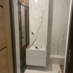 Usługi Wykończeniowe KG - Montaż wiszącej szafki łazienkowej z szufladami, widoczne podłączenia hydrauliczne, białe płytki imitujące marmur na ścianach i podłodze, kabina prysznicowa z czarnymi ramami w tle.