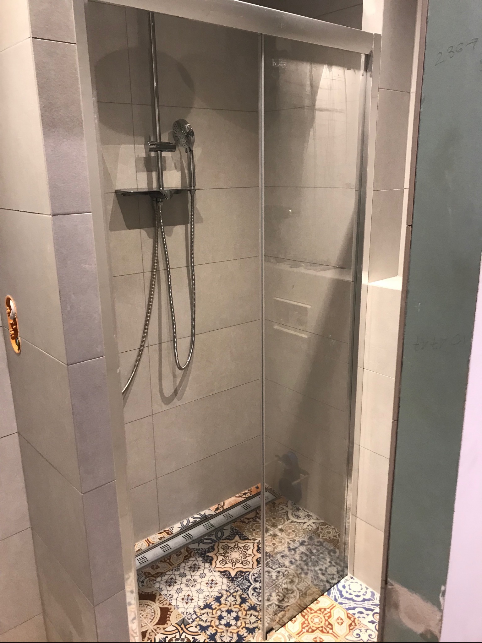 Szklana kabina prysznicowa z chromowaną armaturą na tle szarych płytek ściennych i wzorzystej podłogi. Widoczne niedokończone wykończenia na ścianie po prawej stronie.