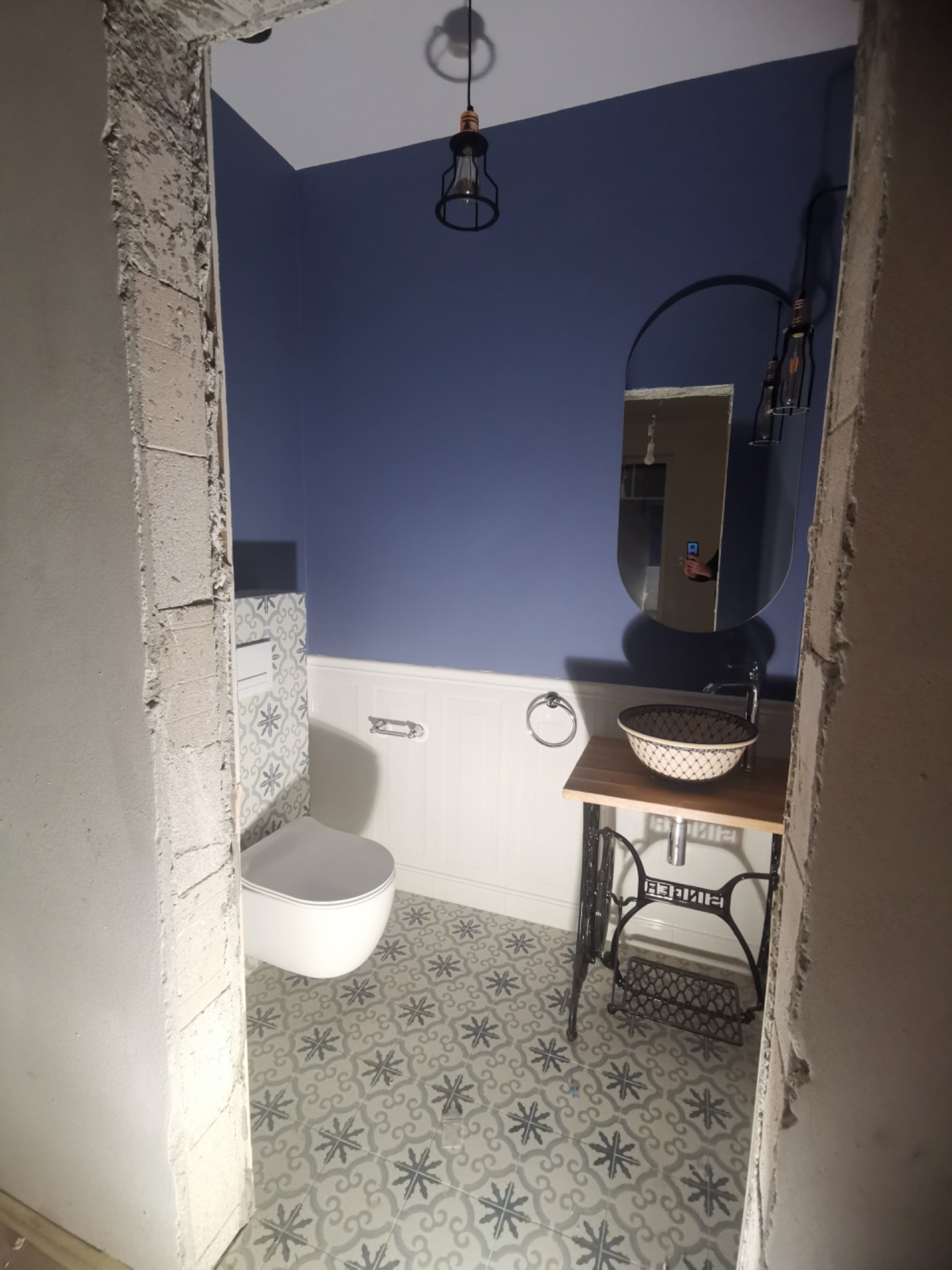 Niebieska ściana w wykończonej łazience z toaletą i umywalką w stylu retro na podstawie maszyny do szycia Singer, widoczne surowe wykończenie otworu drzwiowego.