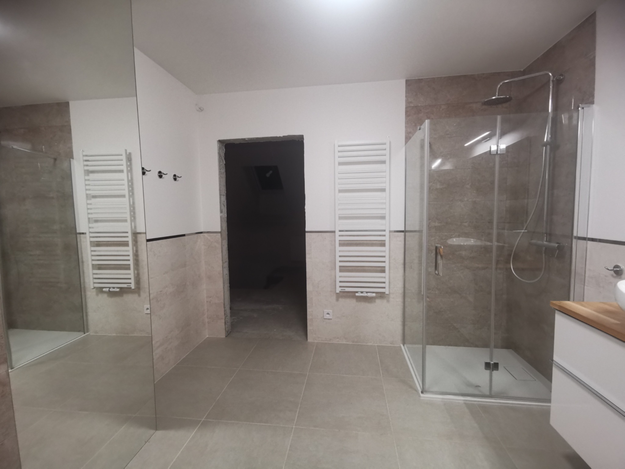 Wyremontowana łazienka z kabiną prysznicową typu walk-in, grzejnikiem drabinkowym, lustrem na ścianie i umywalką z drewnianym blatem; widoczne płytki w odcieniach beżu i brązu oraz surowe wejście...