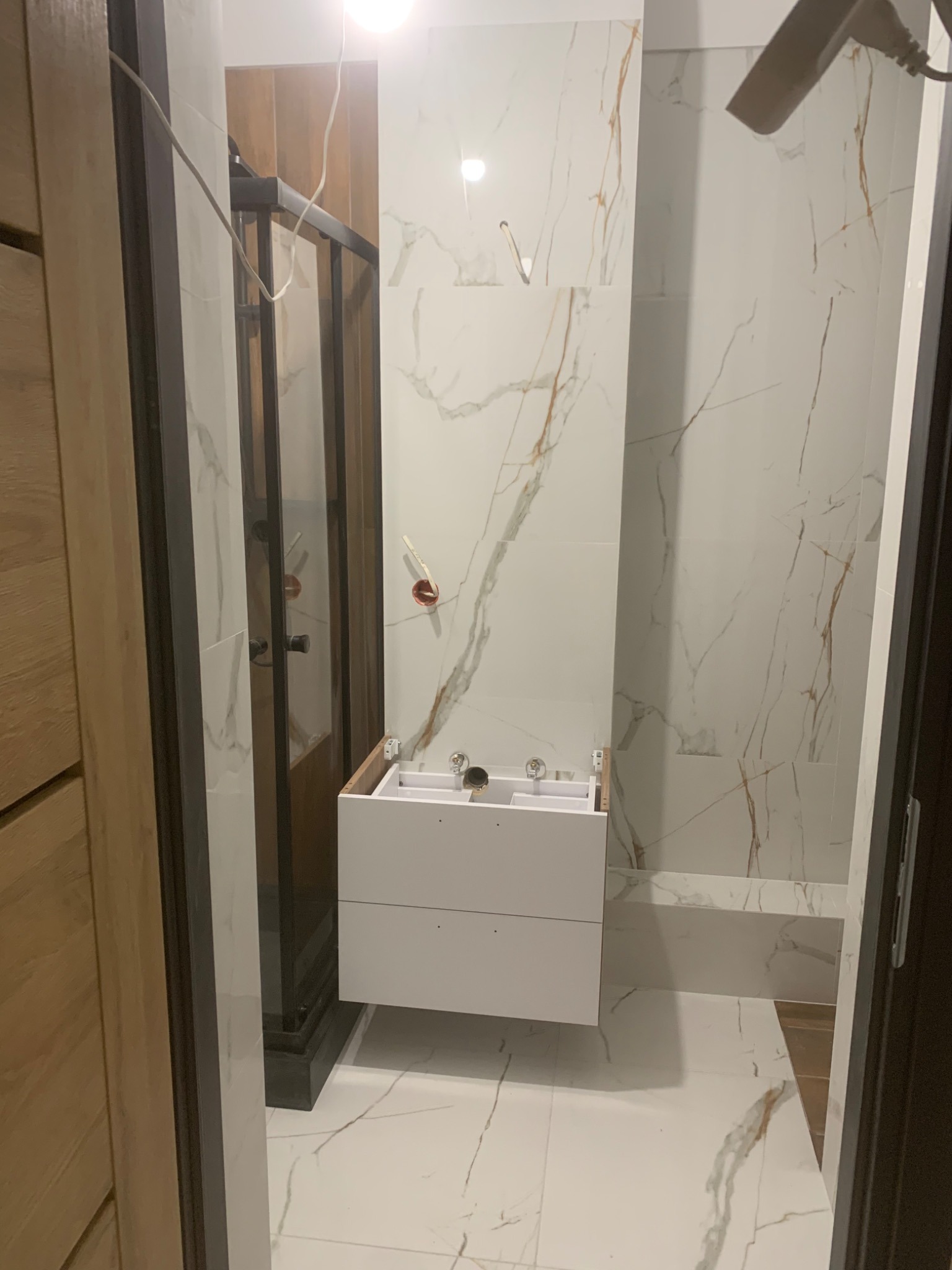 Montaż wiszącej szafki łazienkowej z szufladami, widoczne podłączenia hydrauliczne, białe płytki imitujące marmur na ścianach i podłodze, kabina prysznicowa z czarnymi ramami w tle.