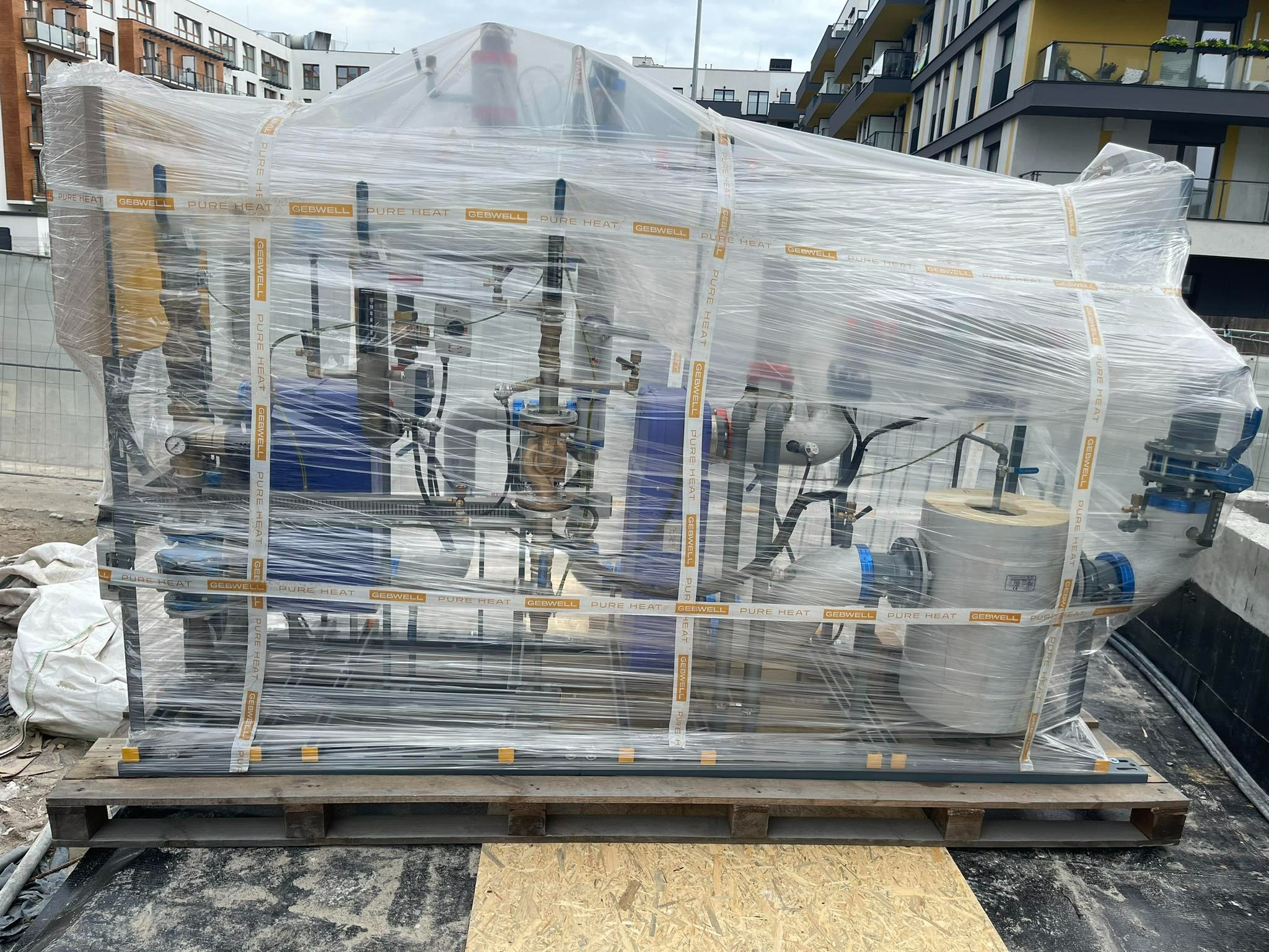 Nowy moduł kotłowni owinięty folią stretch z taśmami GEBWELL PURE HEAT, umieszczony na palecie drewnianej, gotowy do montażu na tle budynków mieszkalnych.