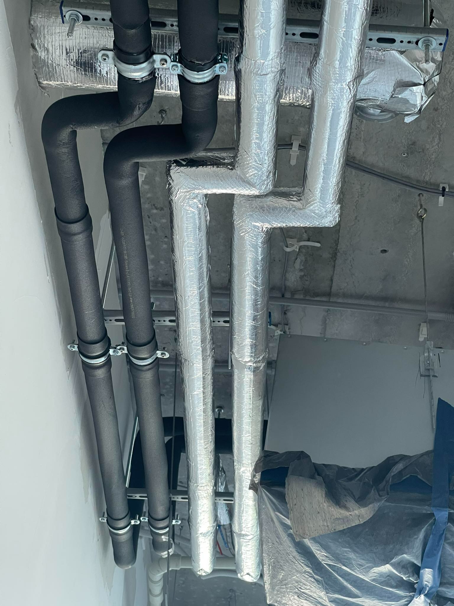 Izolowane rury grzewcze, część czarna, część owinięta srebrną folią, przymocowane do sufitu za pomocą metalowych uchwytów.