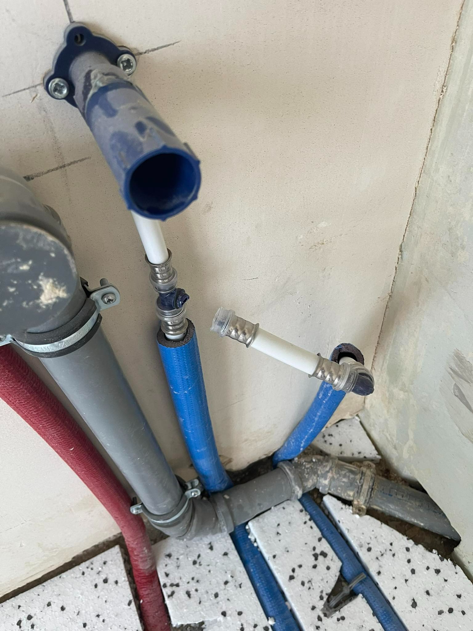 Węzeł instalacji wodnej z rurami w kolorach czerwonym, szarym i niebieskim, częściowo ukryty w warstwie izolacyjnej pod ścianą, widok z góry pod kątem.