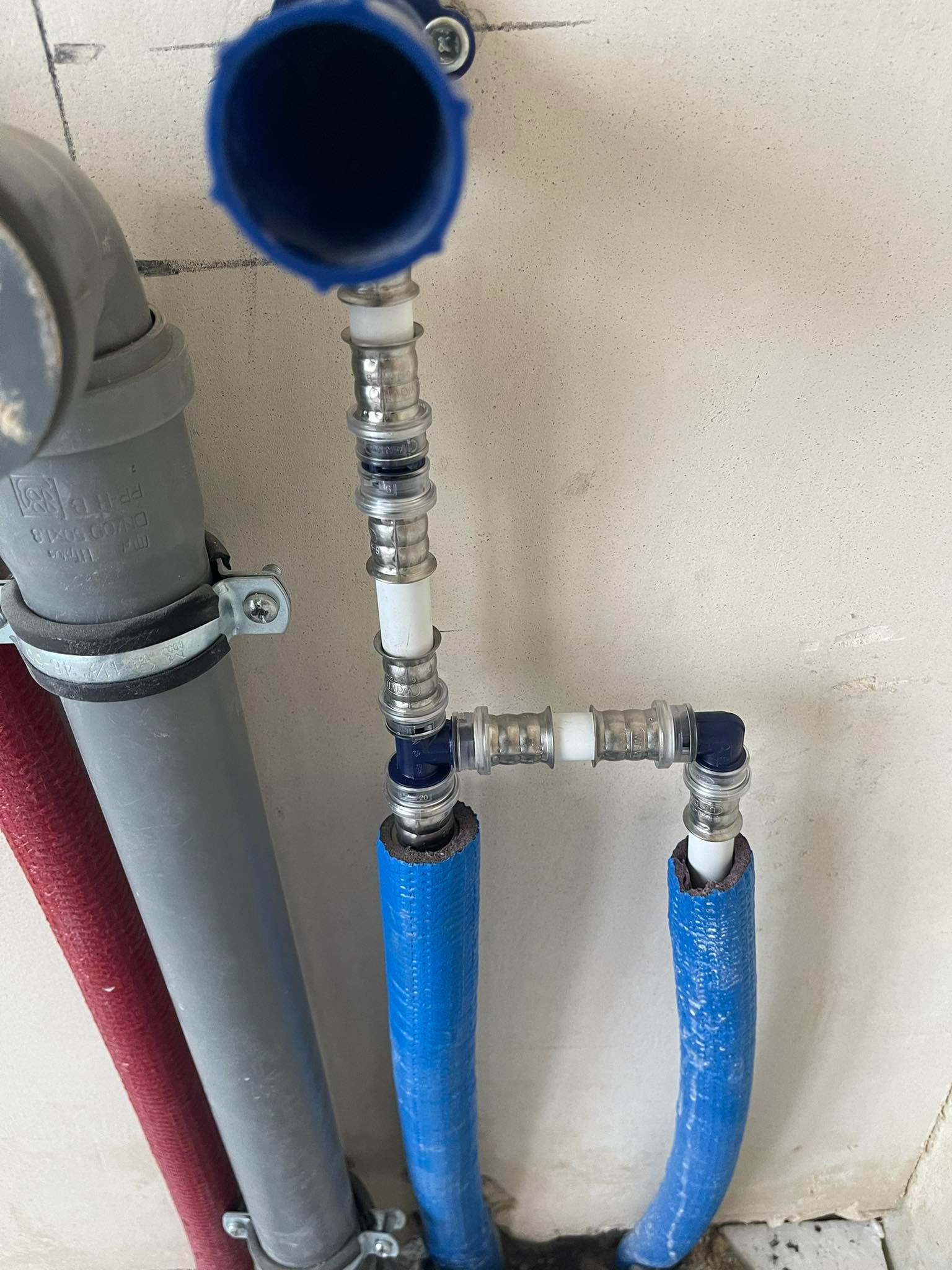 Połączenie rur hydraulicznych z niebieską izolacją termiczną i szarą rurą odpływową zamocowaną do ściany, widoczne złącza zaciskowe.
