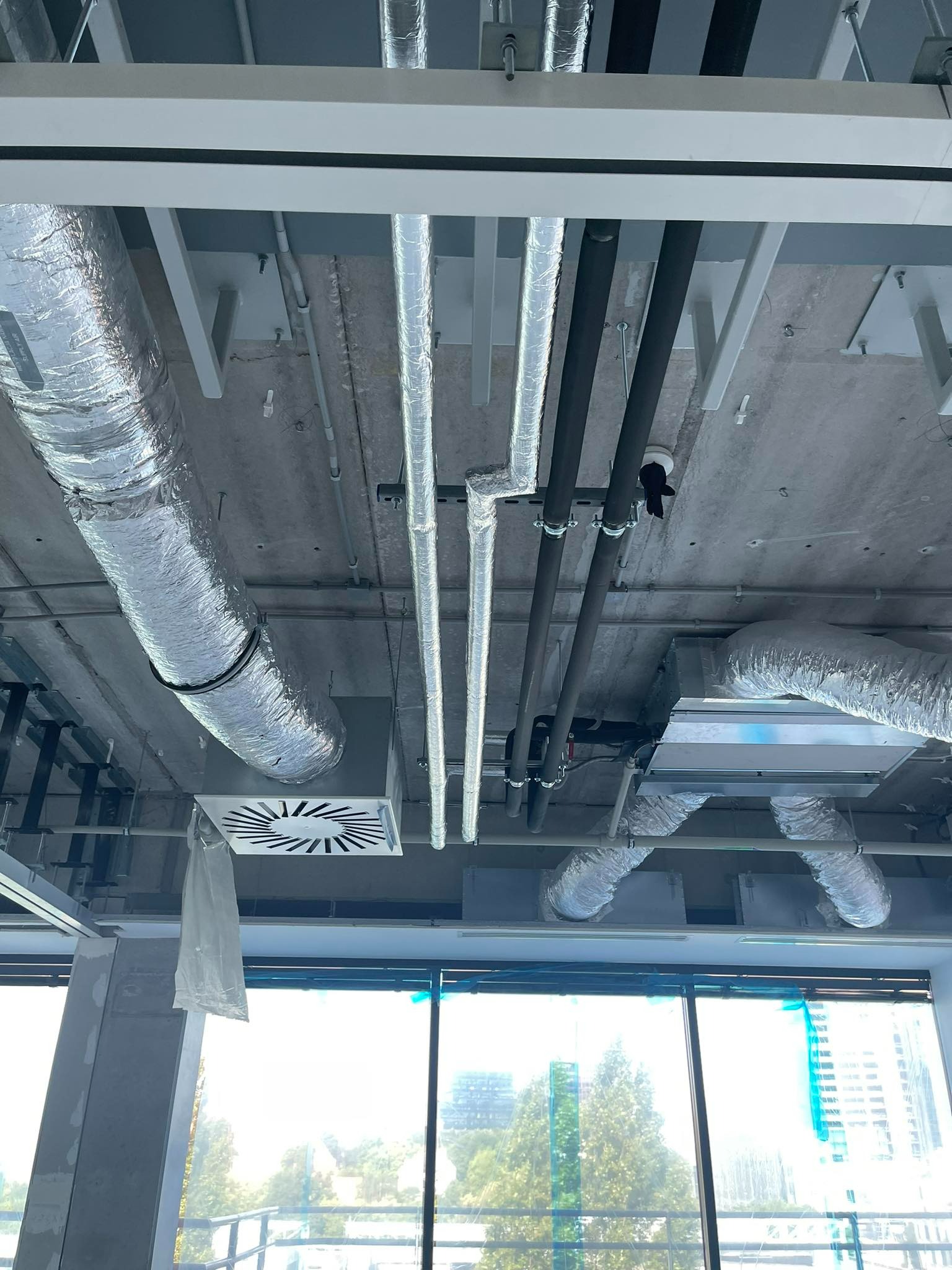 Widok z dołu na sufit w trakcie instalacji systemu wentylacyjnego z widocznymi rurami izolowanymi, czarnymi rurami oraz kratkami wentylacyjnymi, z oknami w tle.