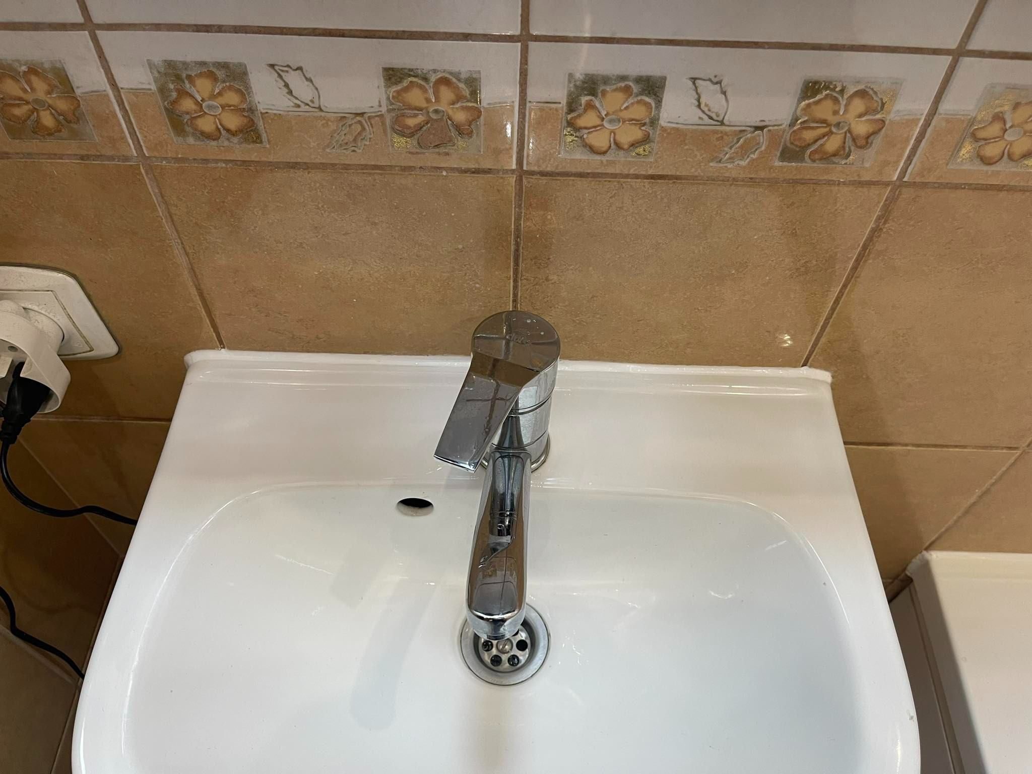 Biała umywalka z chromowaną baterią, brązowe płytki z motywem kwiatowym w tle, widoczny fragment gniazdka elektrycznego.