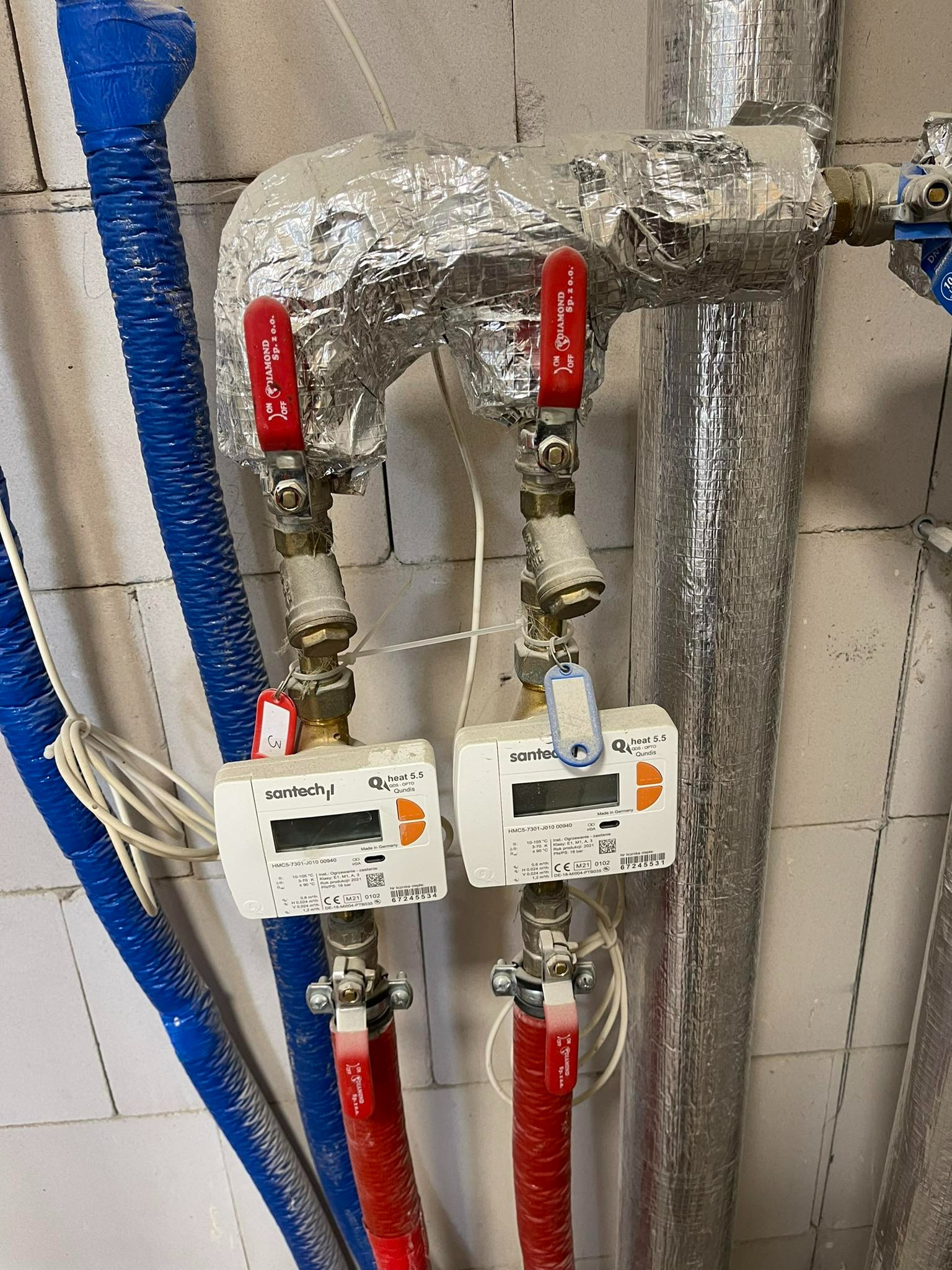 Dwa liczniki ciepła SanTech Q heat 5.5 zamontowane na rurach grzewczych z izolacją aluminiową, z czerwonymi zaworami Diamond Sp. z o.o.