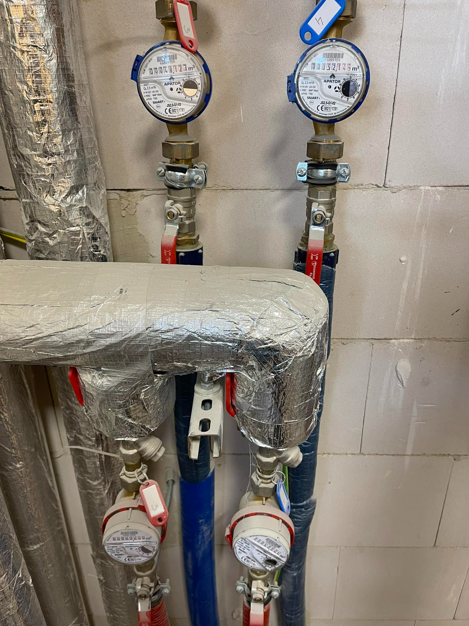 Dwa liczniki wody APATOR SMART+ z zaworami i rurami, zabezpieczone izolacją termiczną, zamontowane na ścianie z bloczków betonowych. Widoczne etykiety z numerami seryjnymi i wskazaniami zużycia.