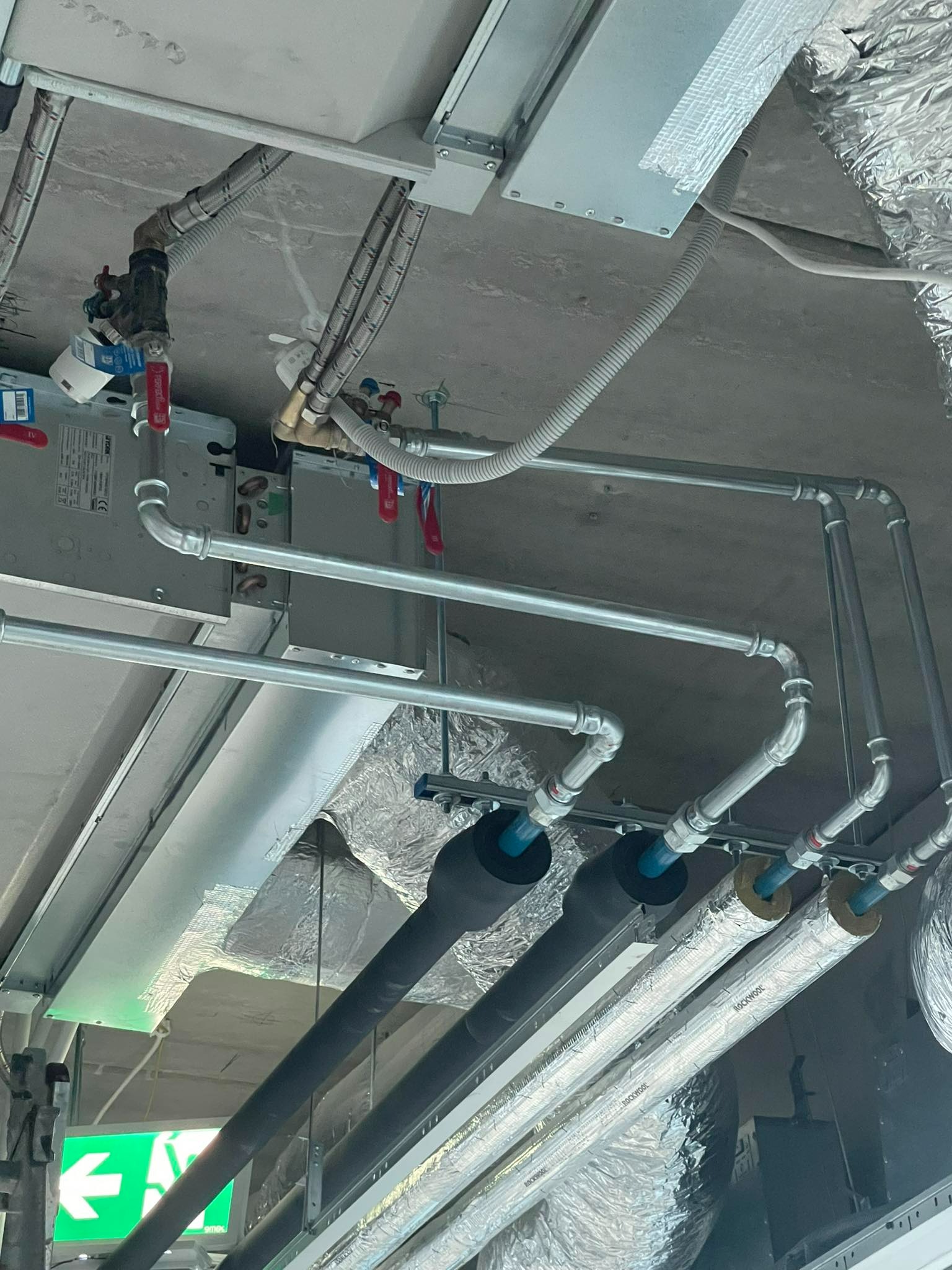 Instalacja rur grzewczych i wodnych pod sufitem, widoczne zawory, izolacja termiczna oraz elementy wentylacji, z uwzględnieniem oświetlenia i oznaczeń bezpieczeństwa.